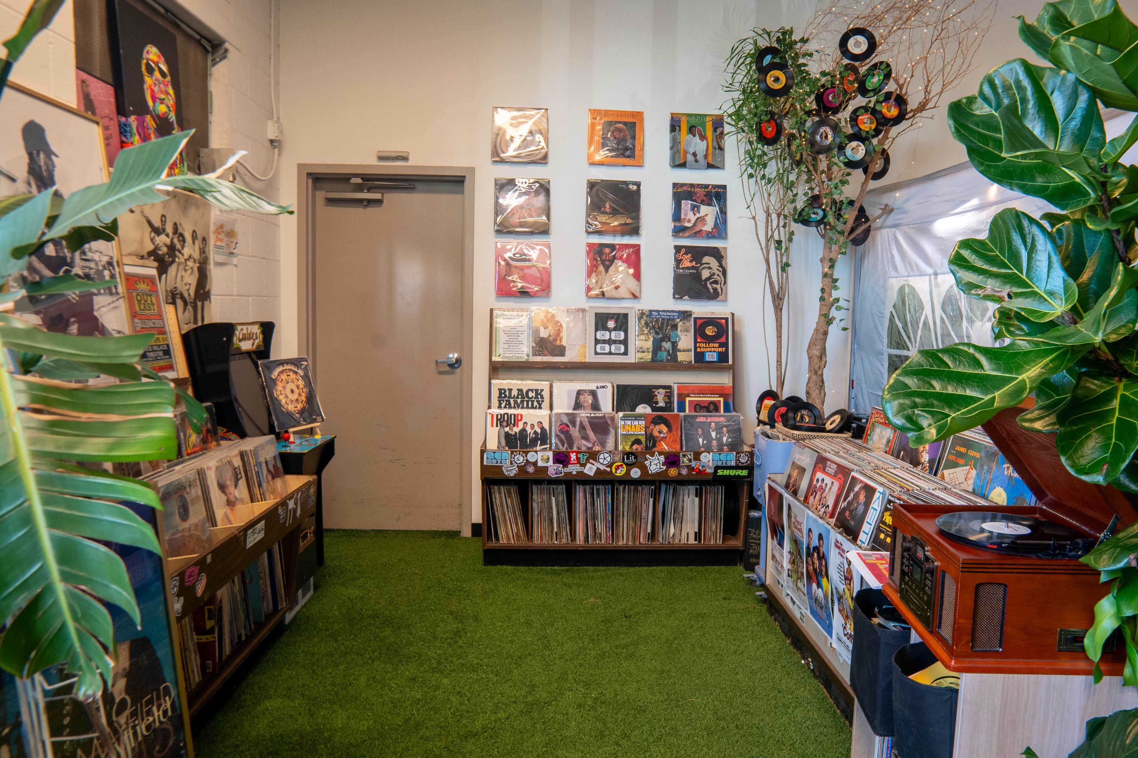 The image shows a vibrant record store with shelves filled with vinyl records, a vintage record player, and decorative plants against a backdrop of colorful album covers on the walls.