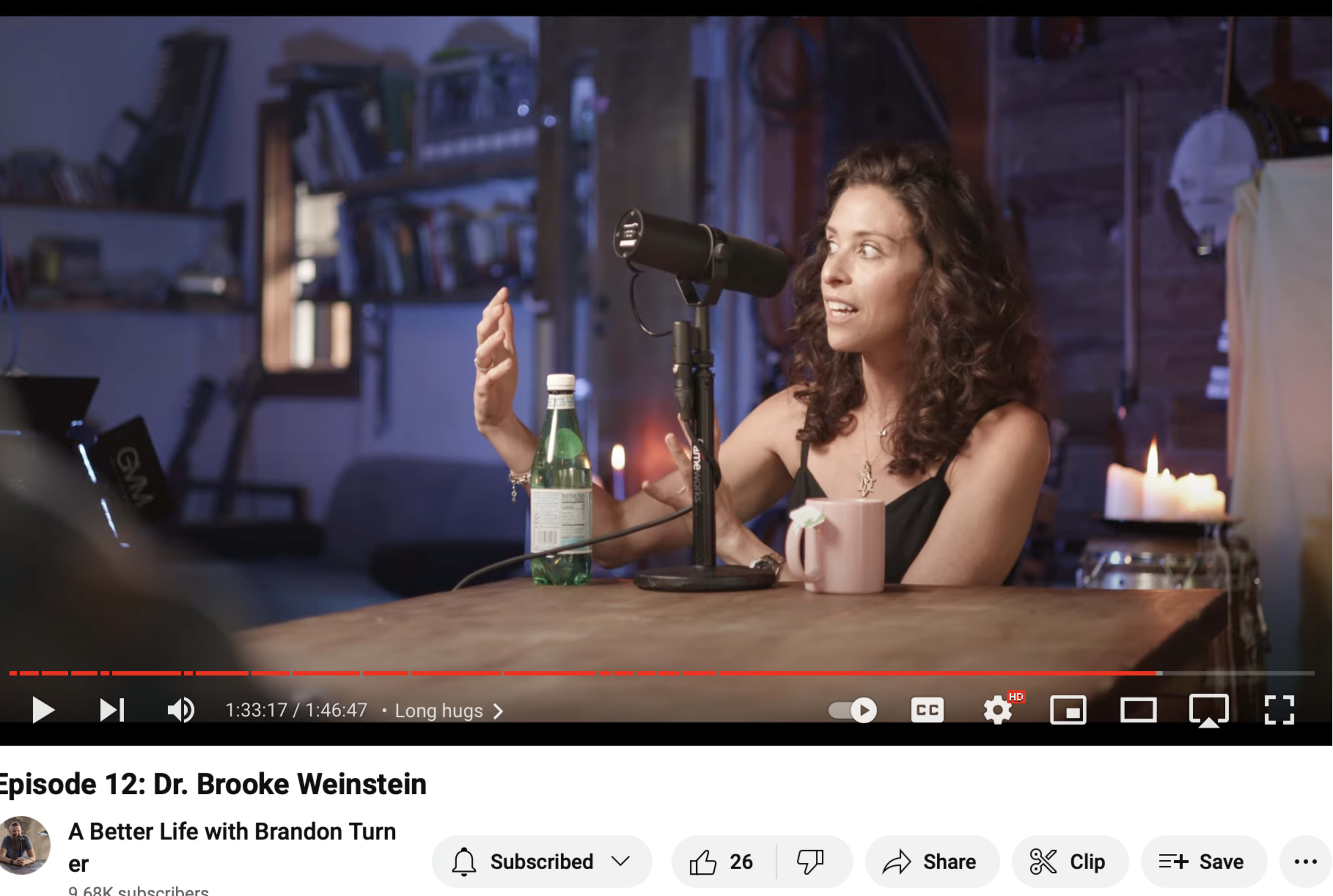 A woman with dark curly hair speaks into a microphone while sitting at a table with a water bottle and a cup.