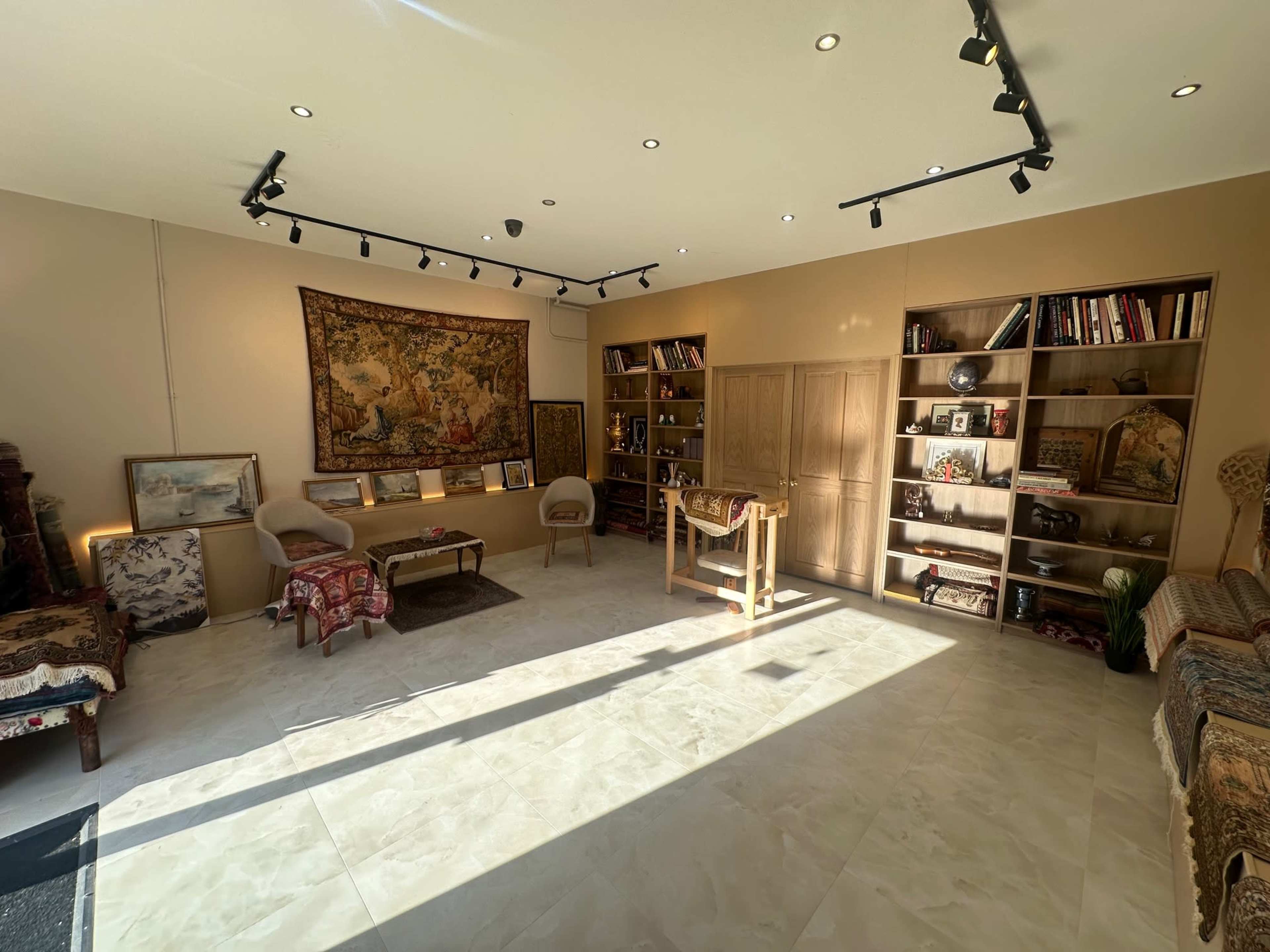 The image shows a well-lit room with wooden shelves filled with books and decorative items, two chairs, a small table, and an ornate tapestry on the wall.