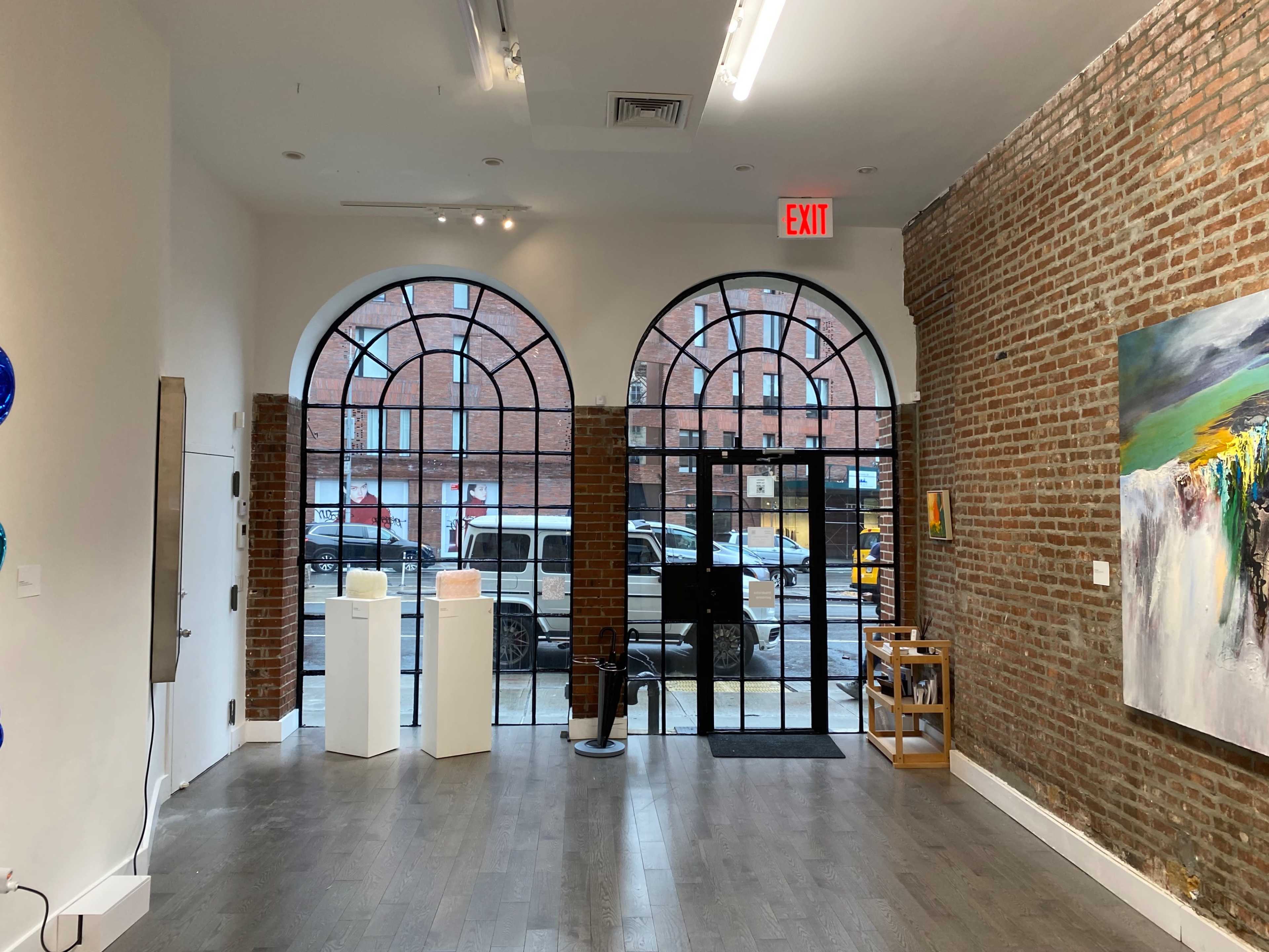 An art gallery interior with large arched windows showcasing a view of the street outside, featuring brick walls, wooden flooring, and a few sculptures displayed on pedestals.