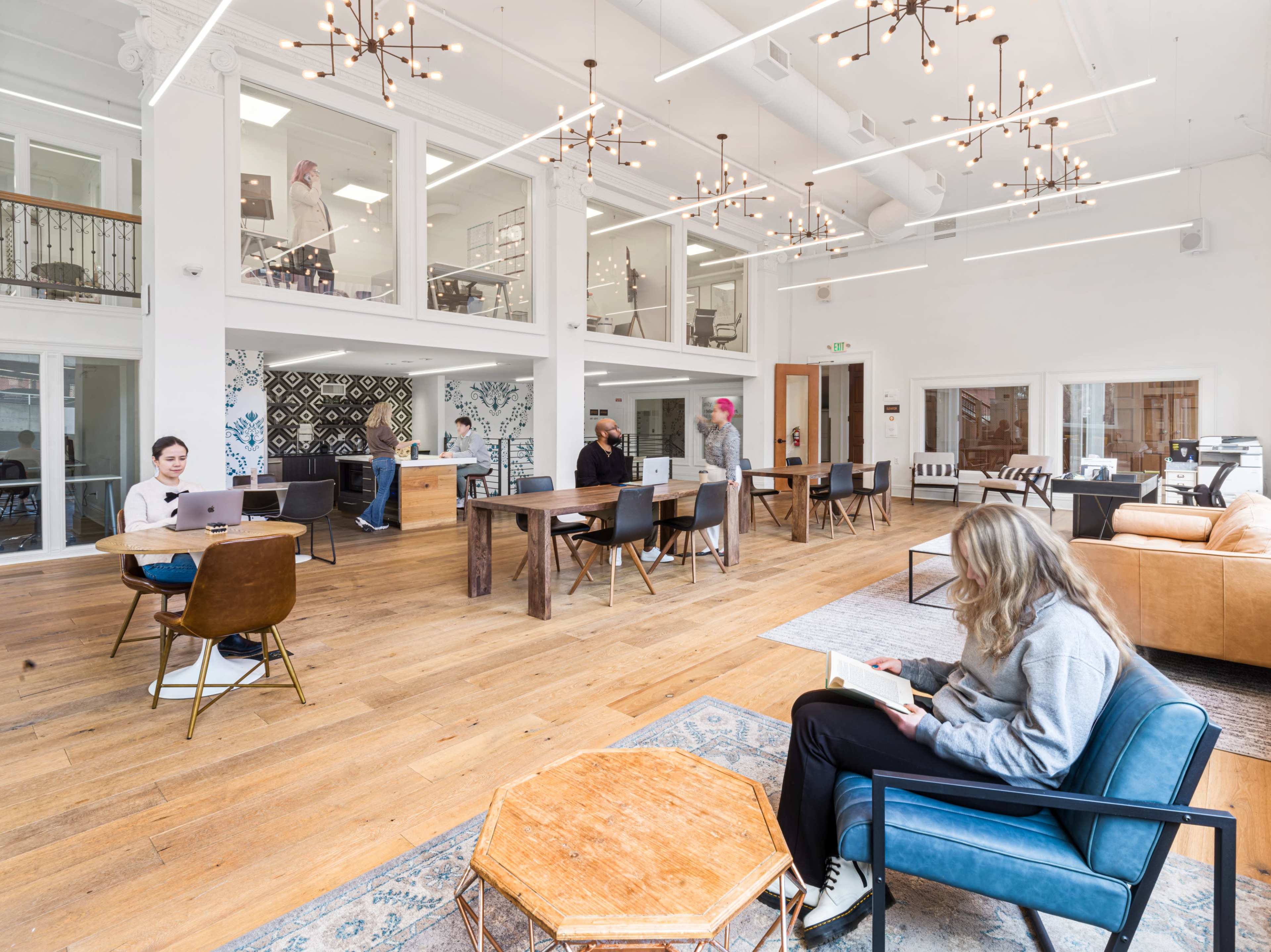 The image shows a modern co-working space with various workstations, seating areas, and large windows, featuring people engaged in different activities.