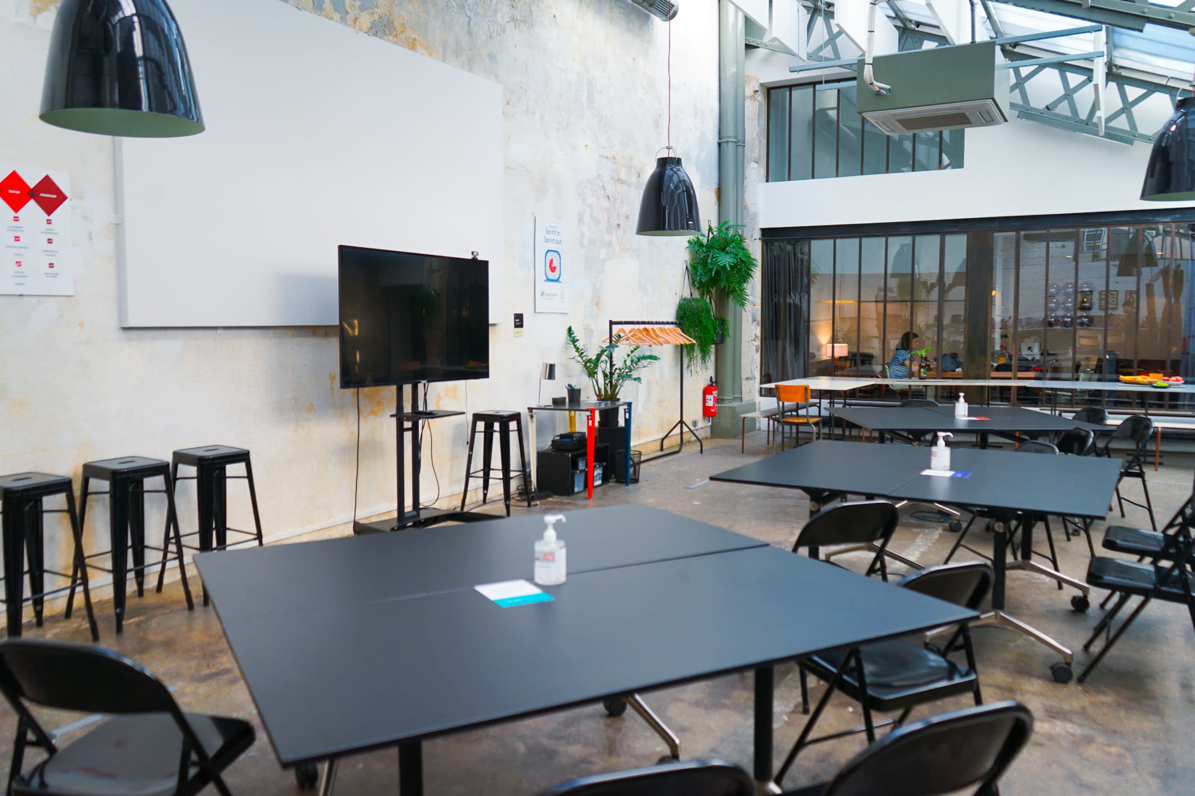 The image shows a modern meeting space with rectangular tables, black chairs, a television on a stand, and large windows in a well-lit industrial room.