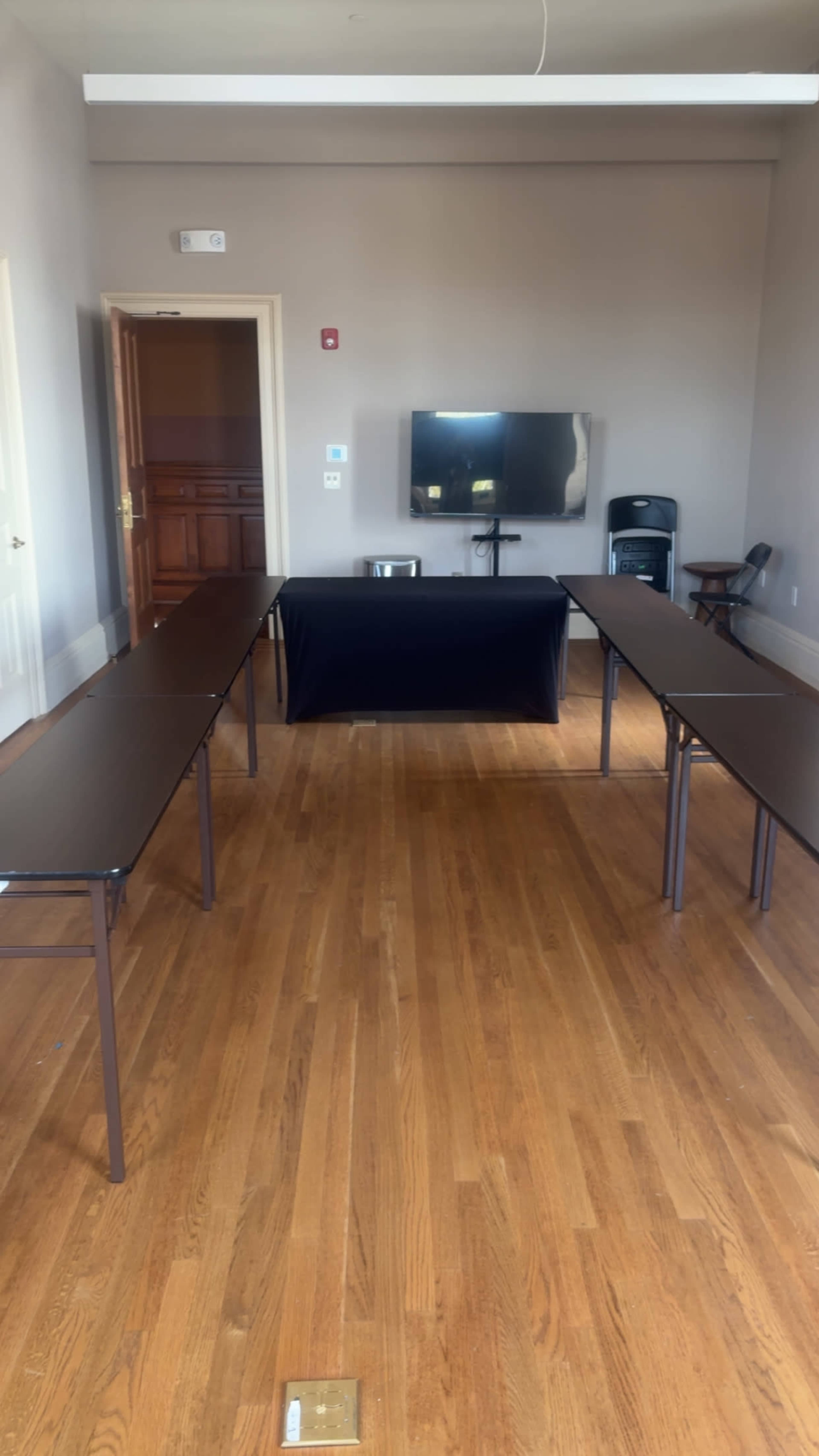 The image shows a room with wooden floors, two long tables arranged in an inverted U-shape, a black tablecloth on a central table, and a wall-mounted TV in the background.