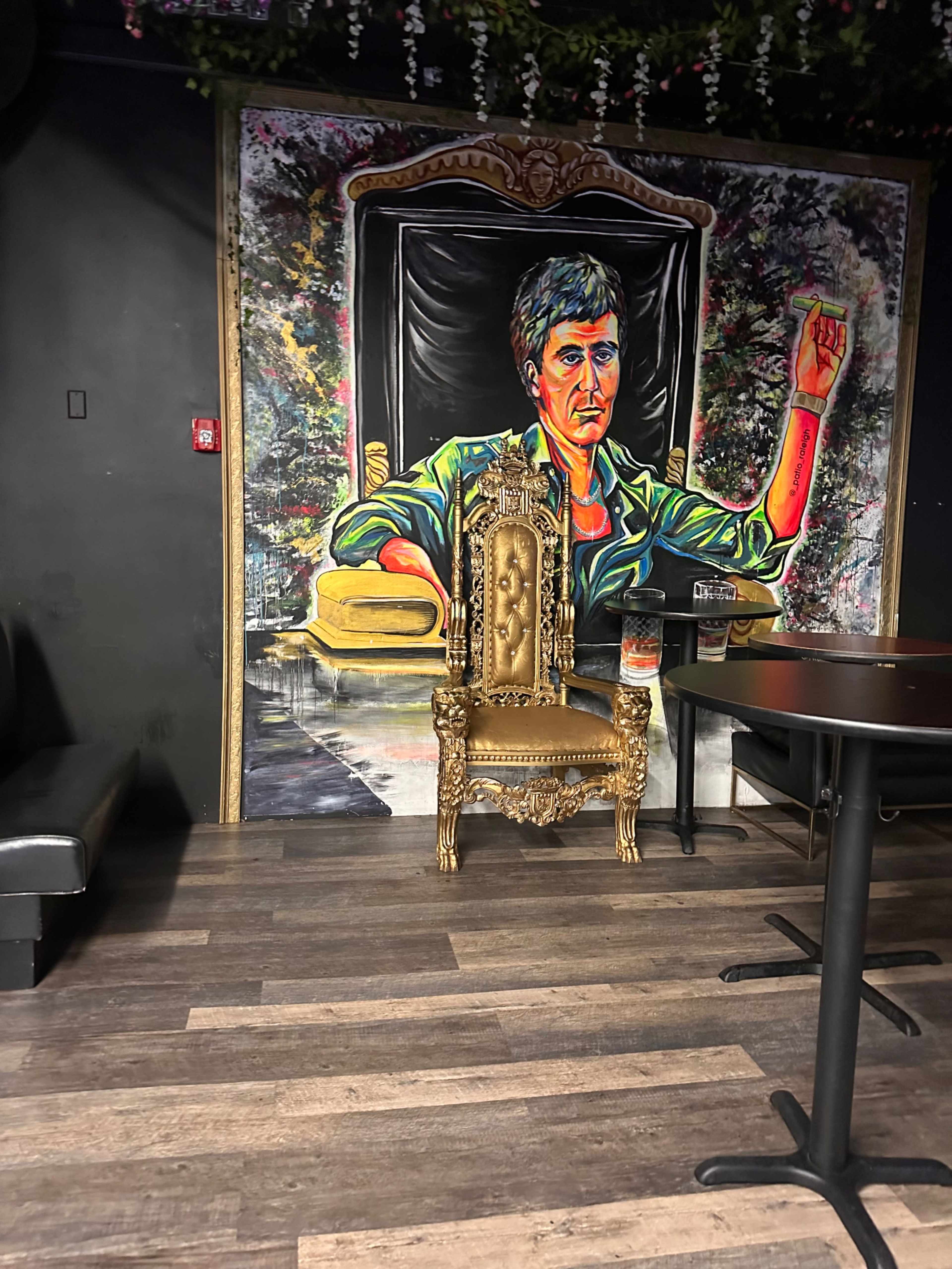 A gold ornate chair in front of a colorful mural depicting a person with short hair and an arm raised, set in a dimly lit room with dark walls and wooden flooring.