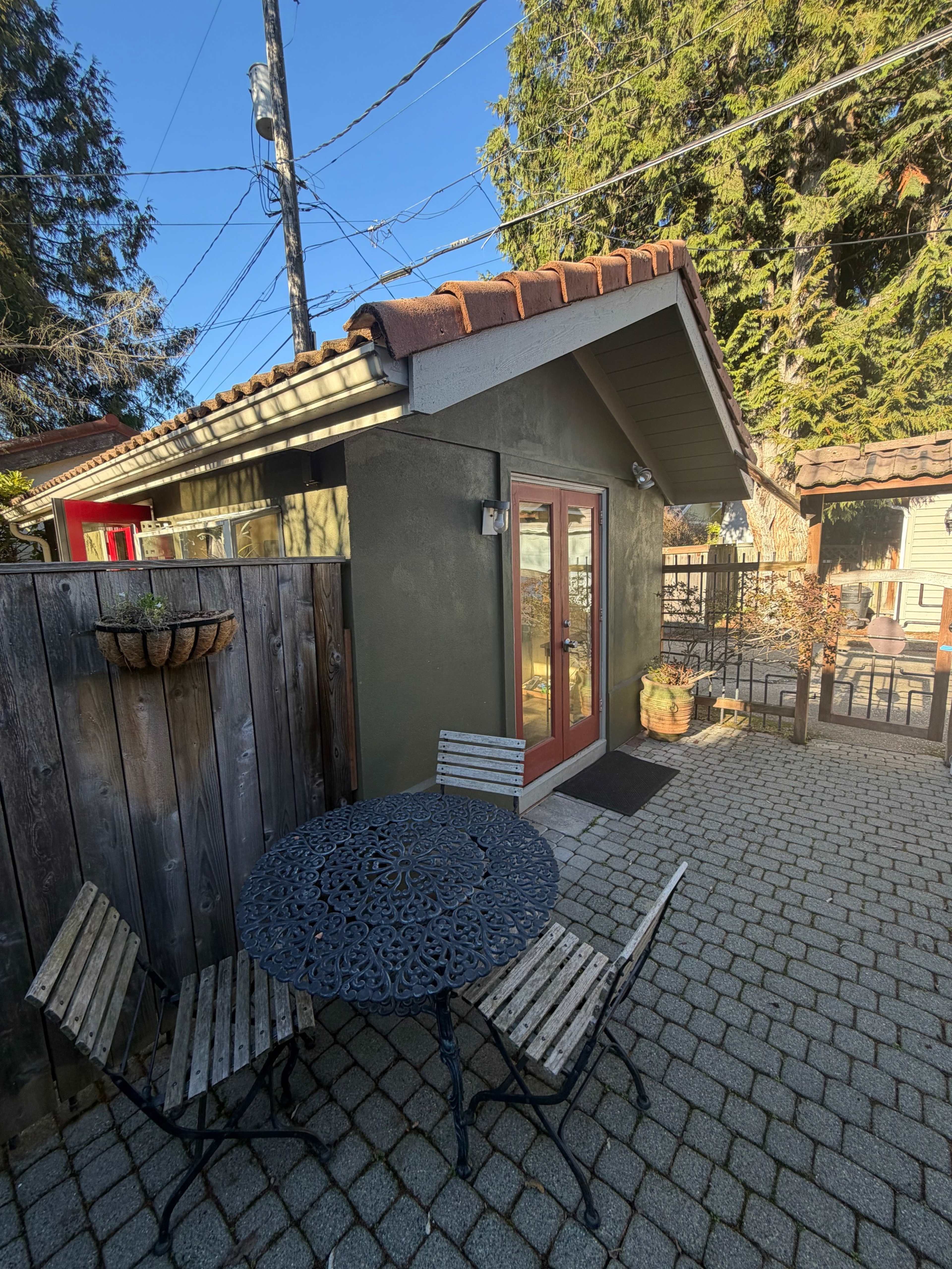 A small outdoor patio features a round metal table and two chairs, adjacent to a green building with red-framed doors, surrounded by a fence and trees.