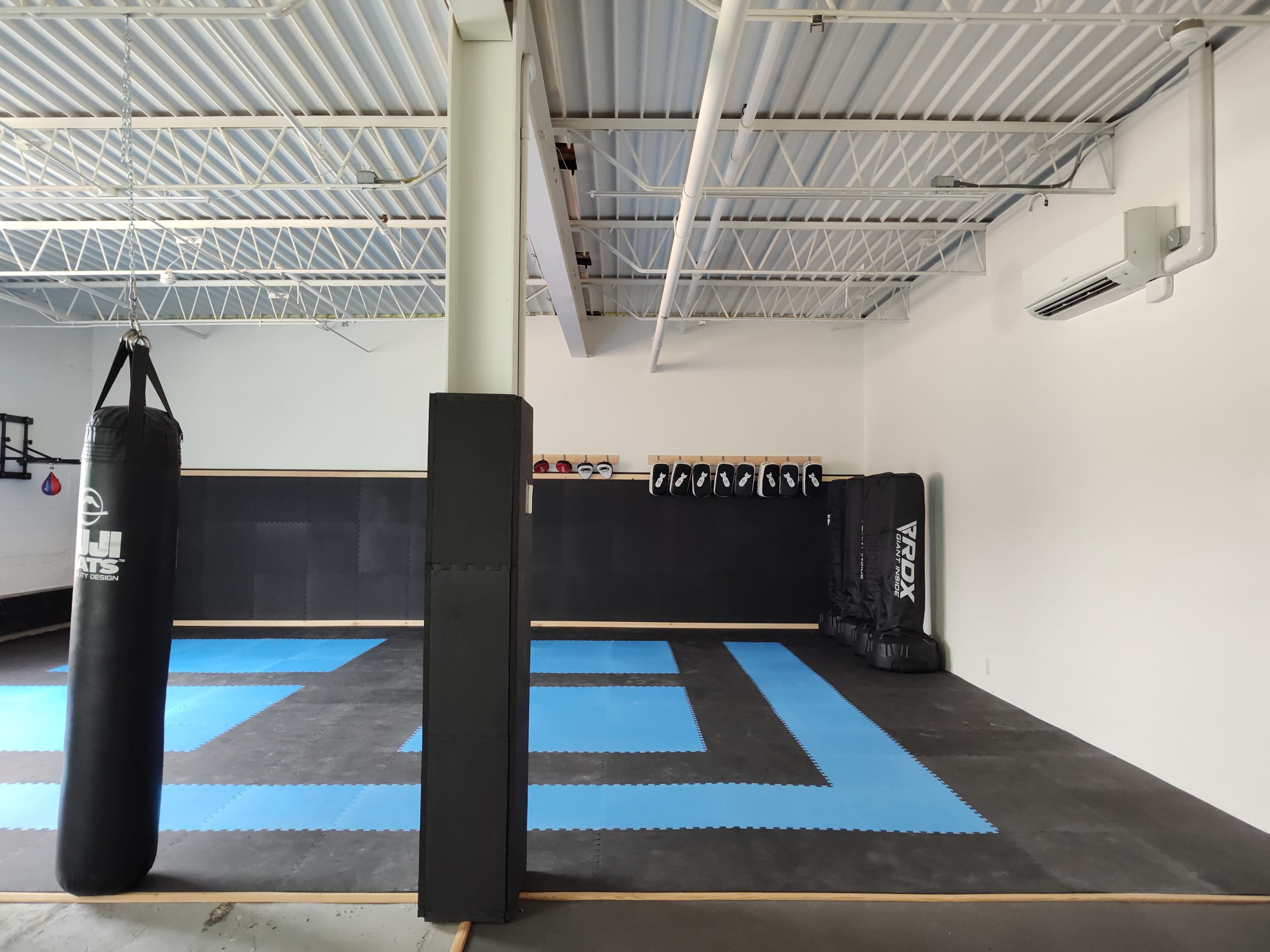 The image shows an empty martial arts training gym featuring a heavy bag, padded walls, and a blue tile floor arranged in a geometric pattern.
