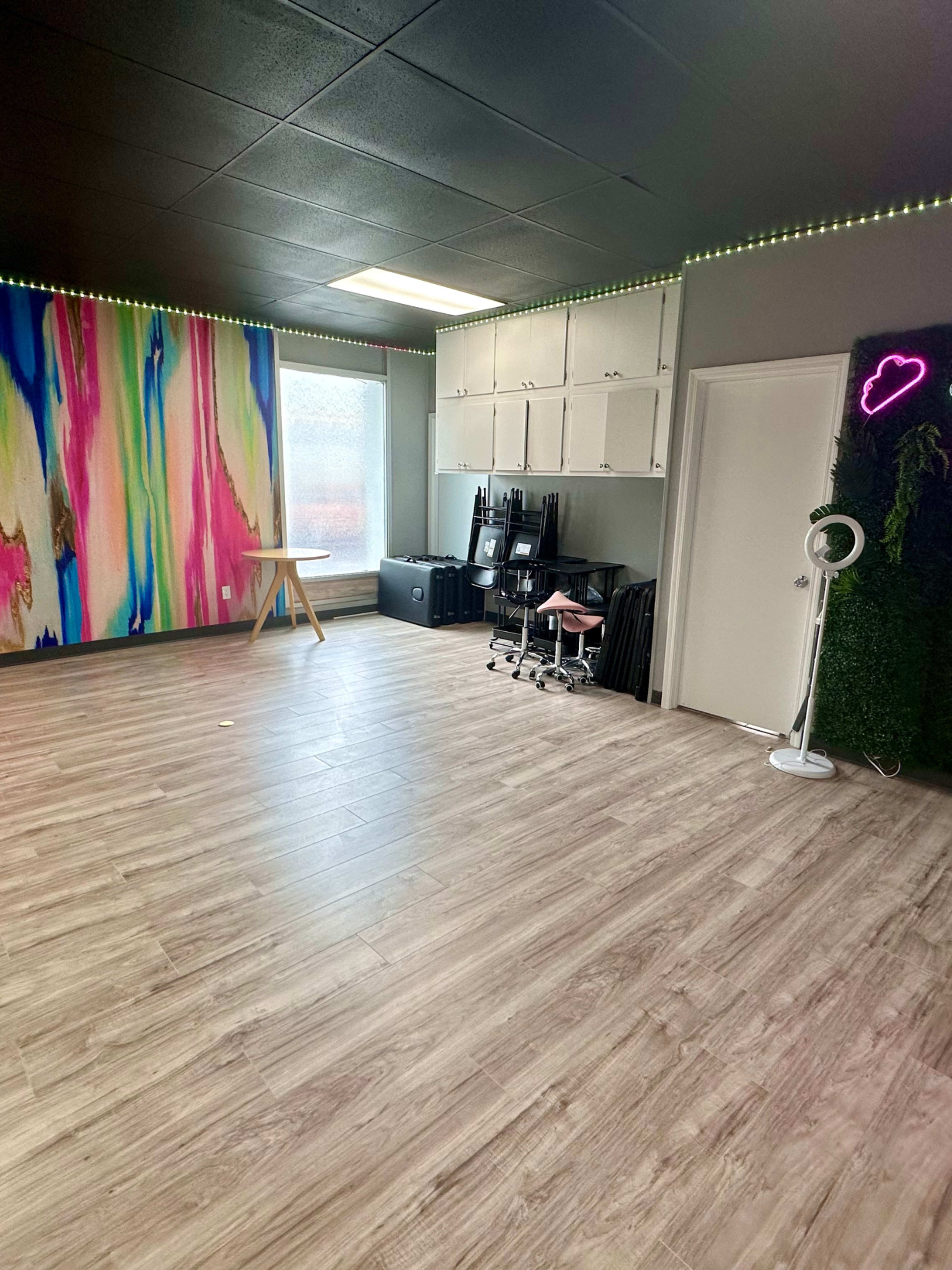 A spacious room with a colorful abstract mural on one wall, wooden flooring, and a setup that includes chairs, a table, and storage cabinets.