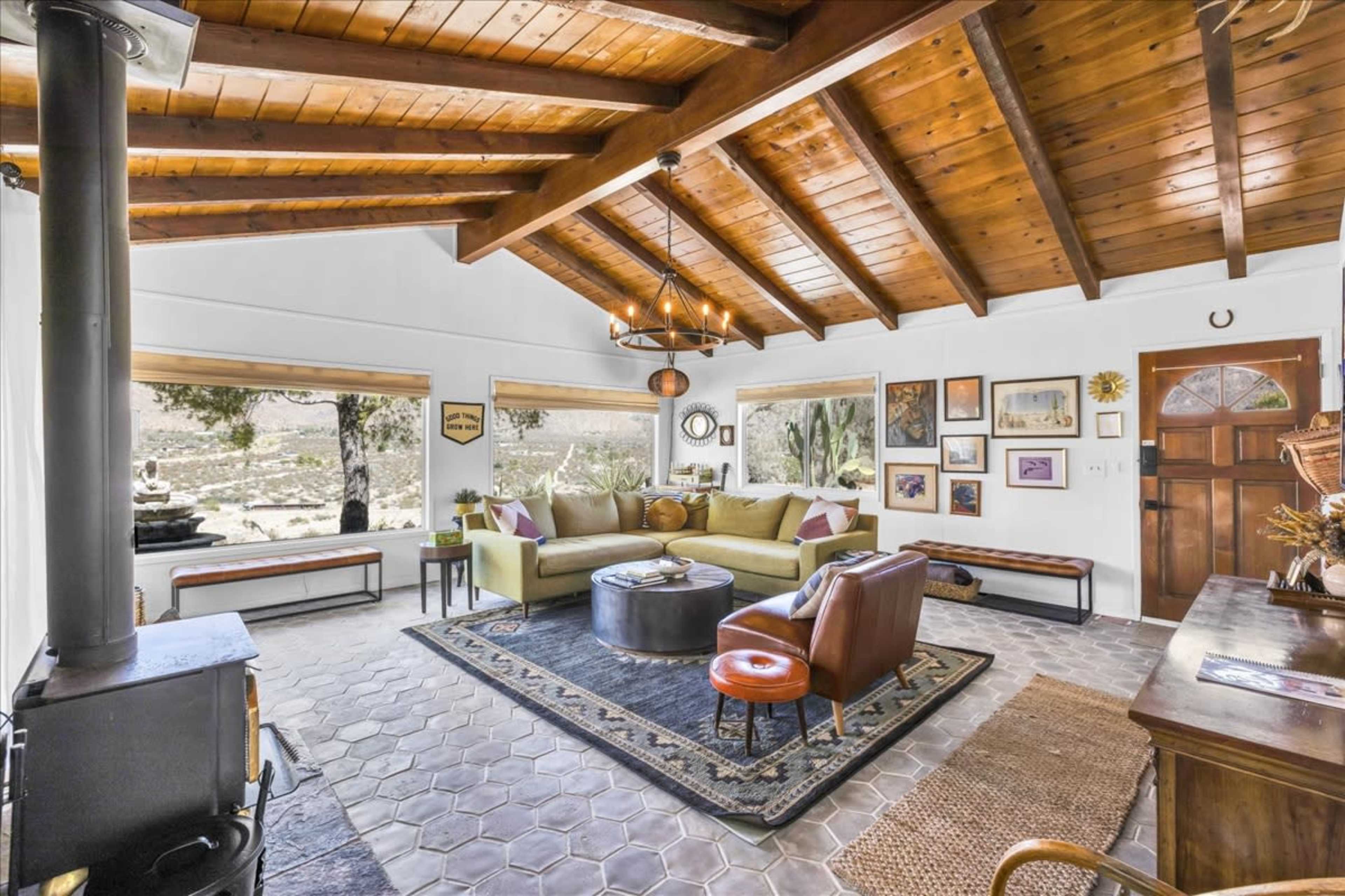 The image shows a cozy living room with a wooden beamed ceiling, a circular coffee table, two chairs, and various artworks on the walls.