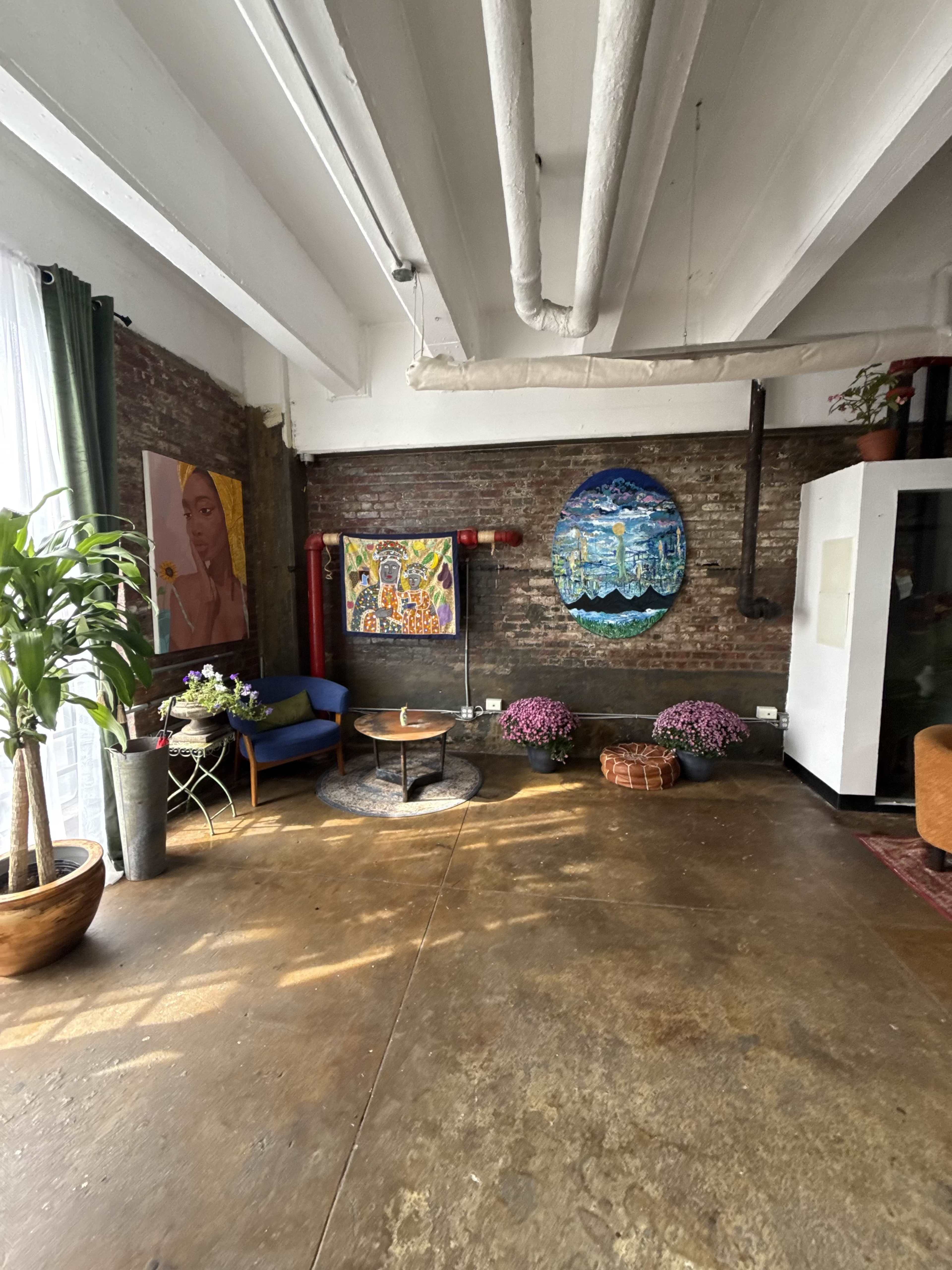 The image shows a spacious, well-lit room with artwork on the walls, plants, and a mix of seating areas.