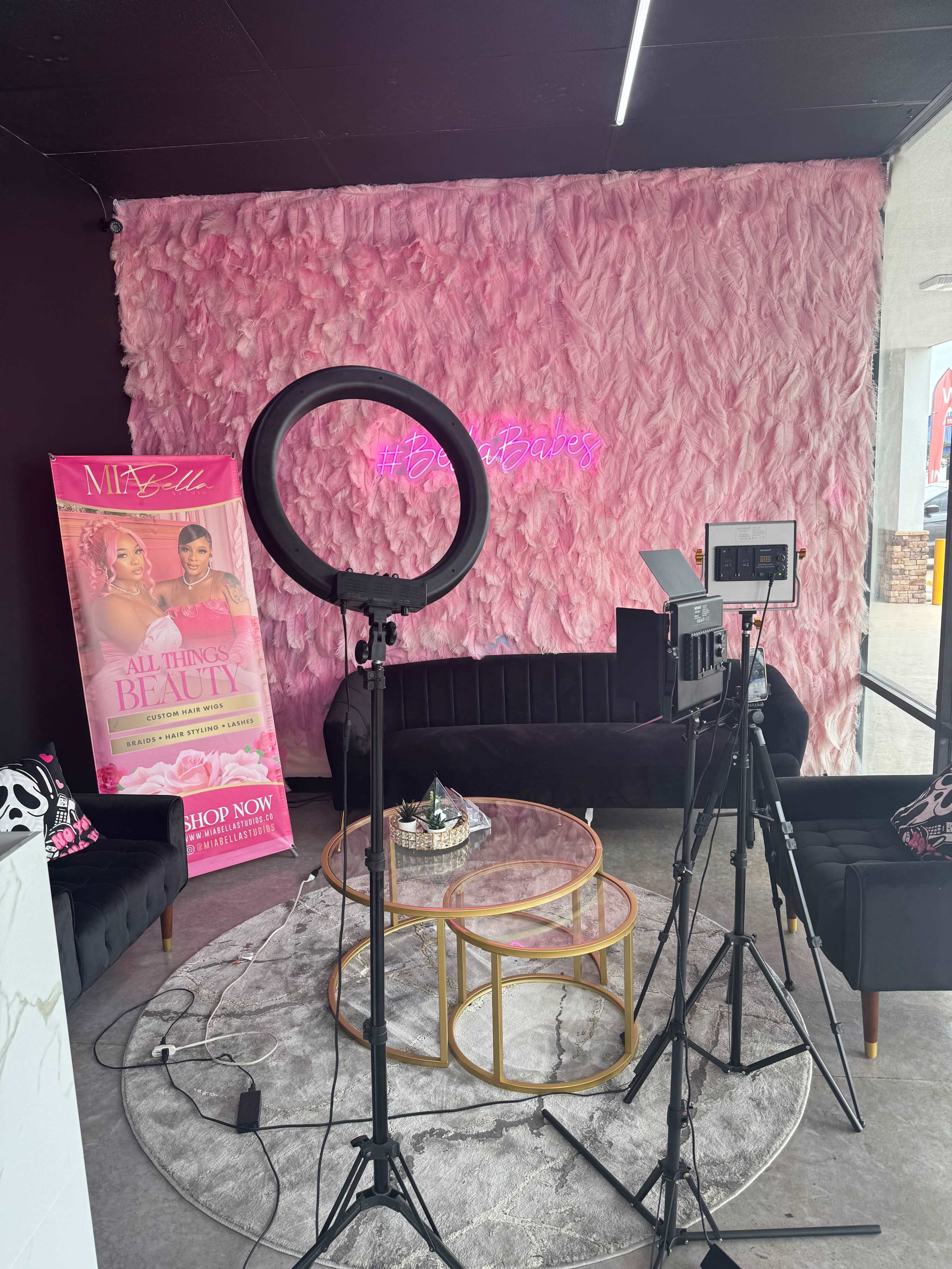A beauty studio features a pink textured wall, a neon sign, a ring light, a camera setup, and a round coffee table with decor items.