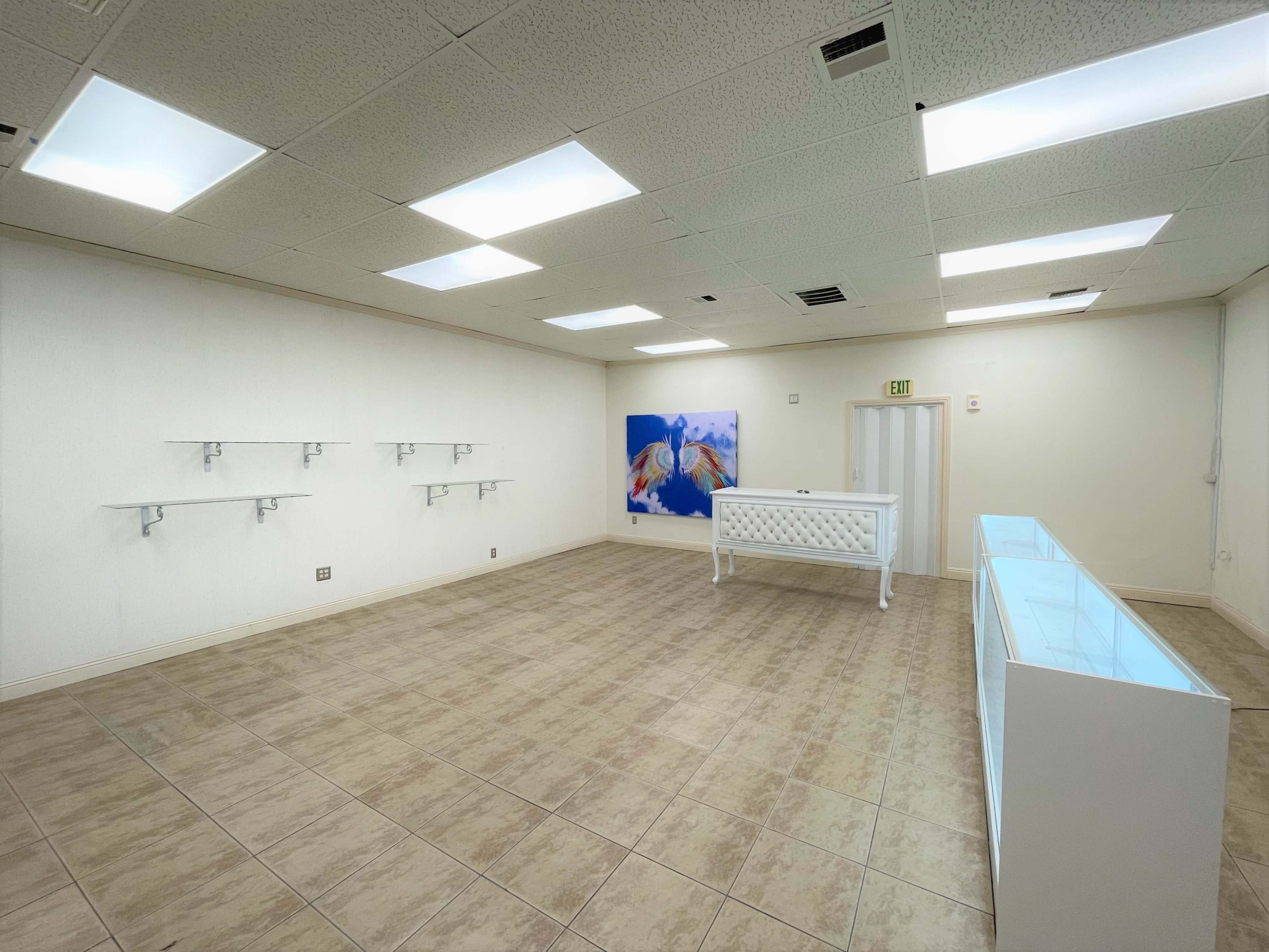 The interior of a vacant retail space features white walls, tiled flooring, a pink display case at the center, and minimal shelving on the walls.