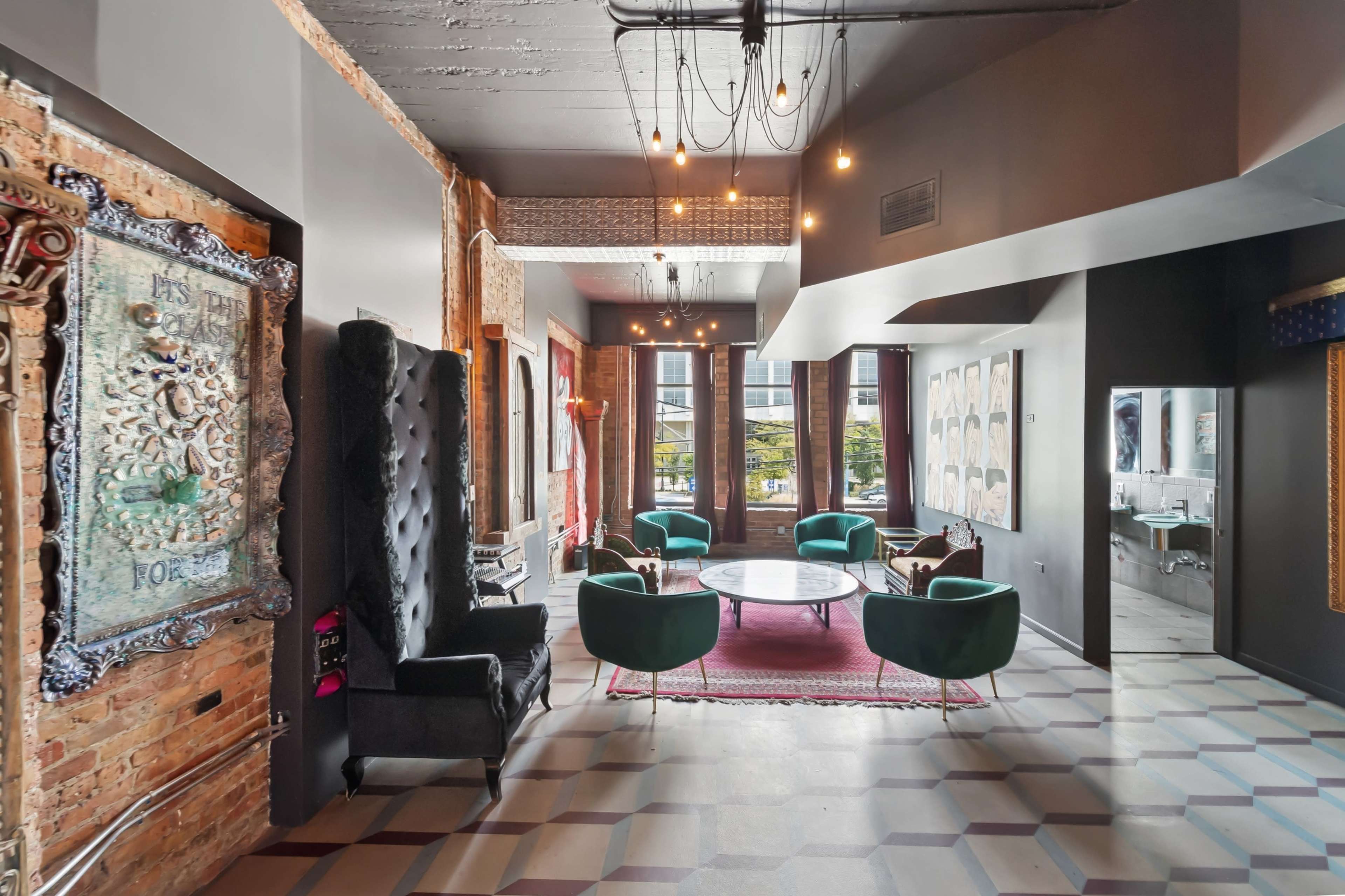 The image shows a modern lounge area with a mix of textured brick walls and sleek decor, featuring green upholstered chairs, a round table, and artistic wall hangings.