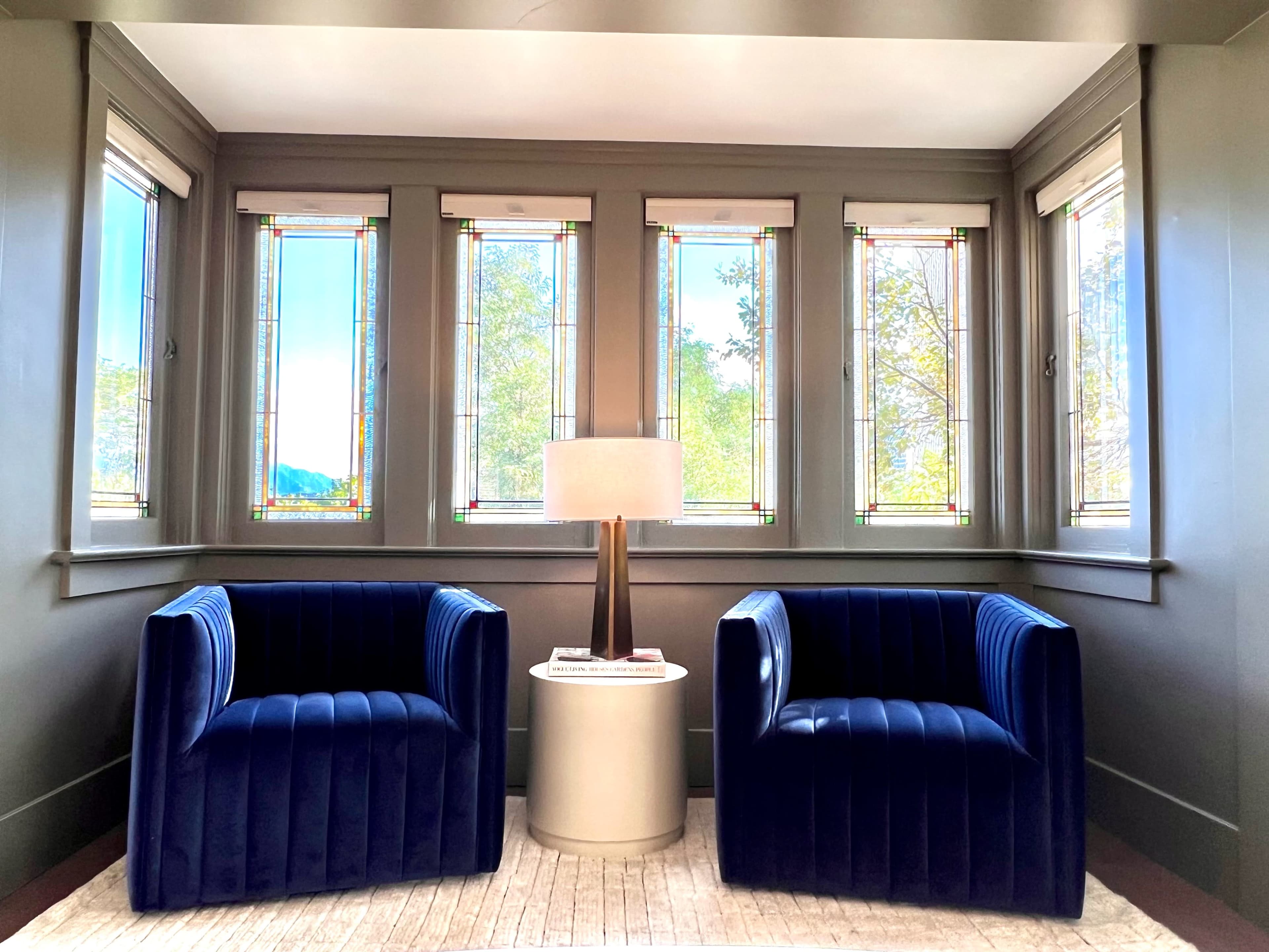A cozy corner of a room featuring two blue upholstered chairs, a round white table, and bright windows with stained glass accents.
