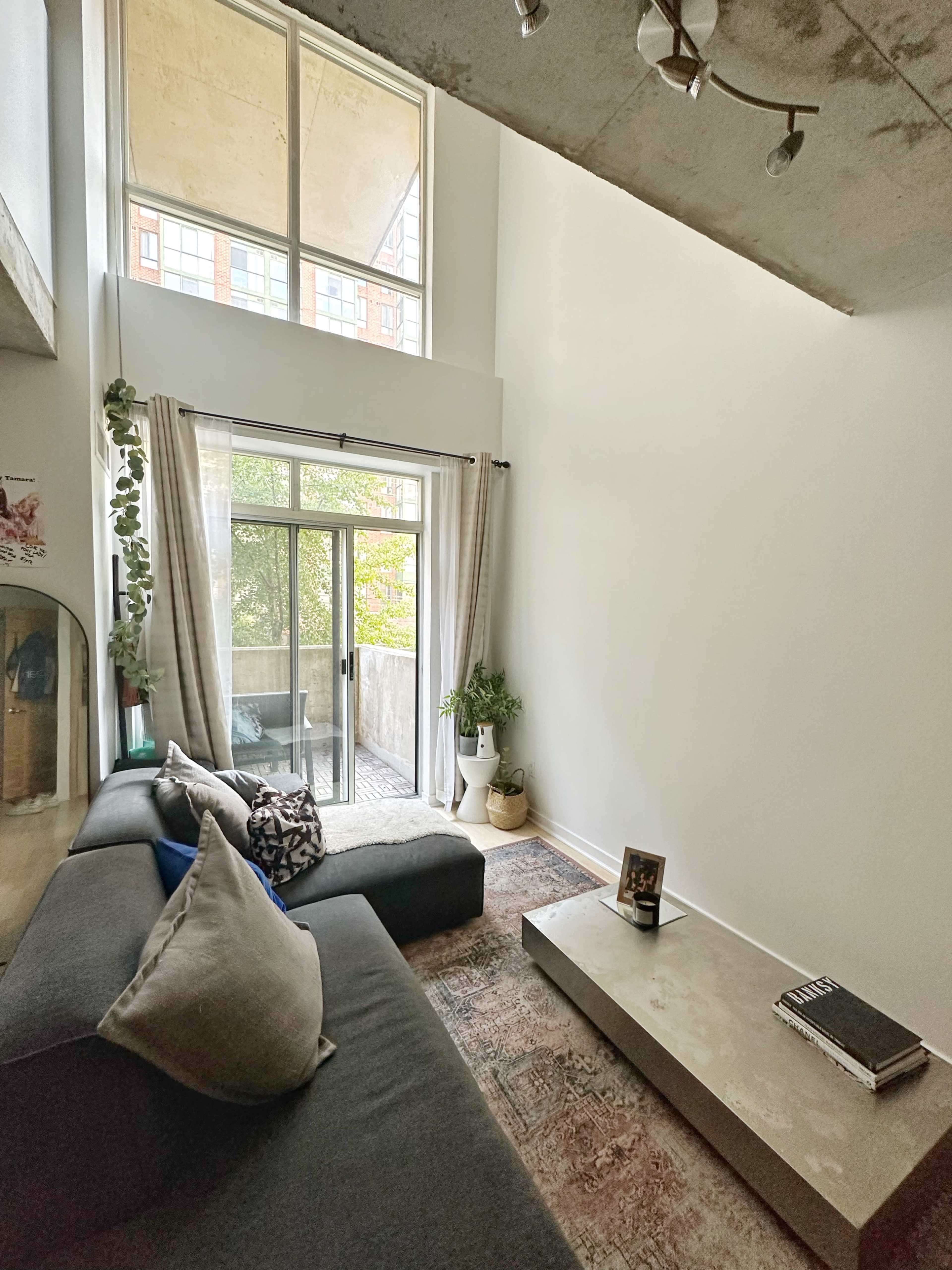 A modern living room with a black sofa, a concrete coffee table, large windows, and a balcony overlooking greenery.