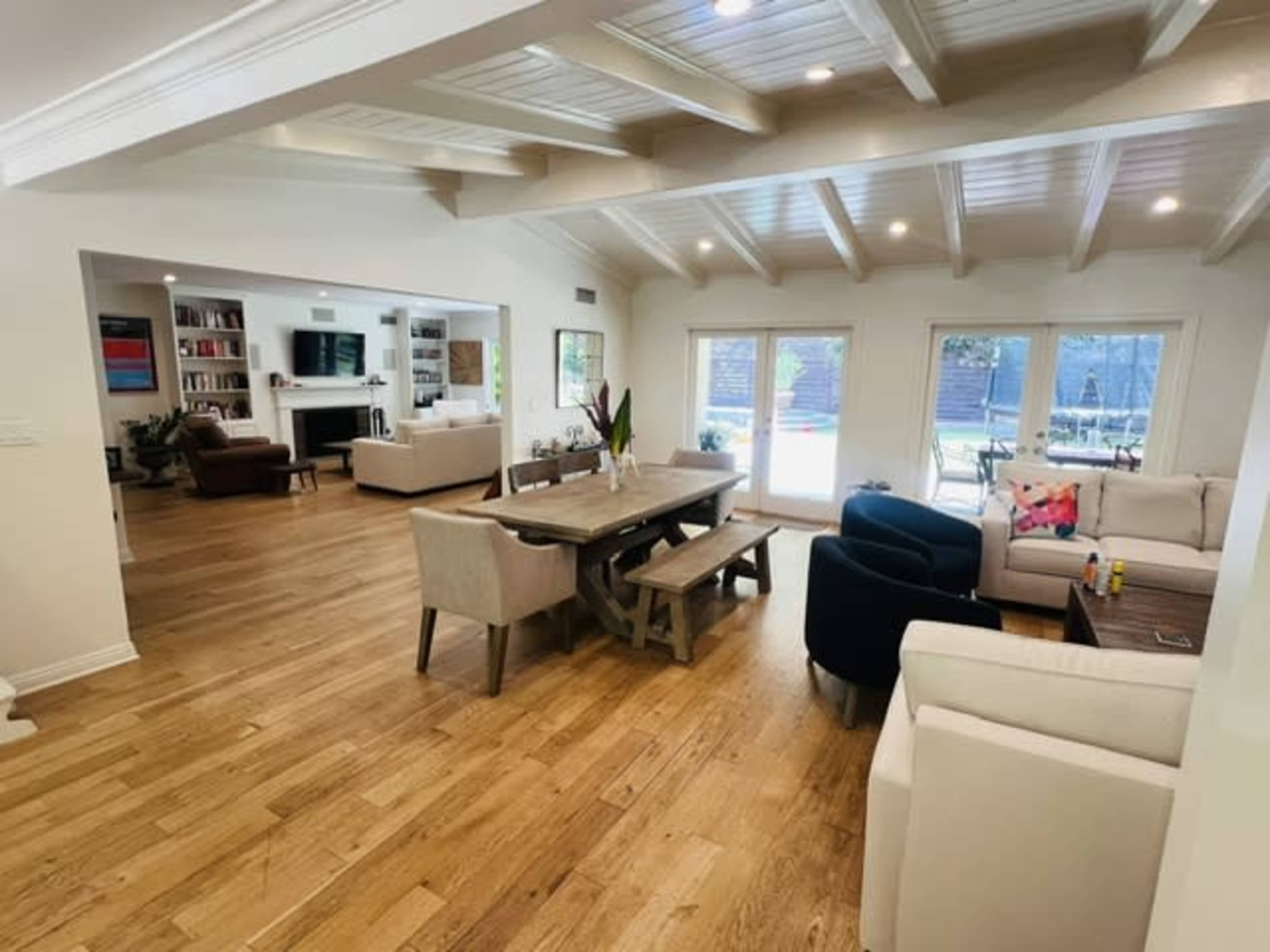 The image shows an open-concept living space with a wooden dining table, comfortable seating areas, and large windows overlooking a pool in the backyard.