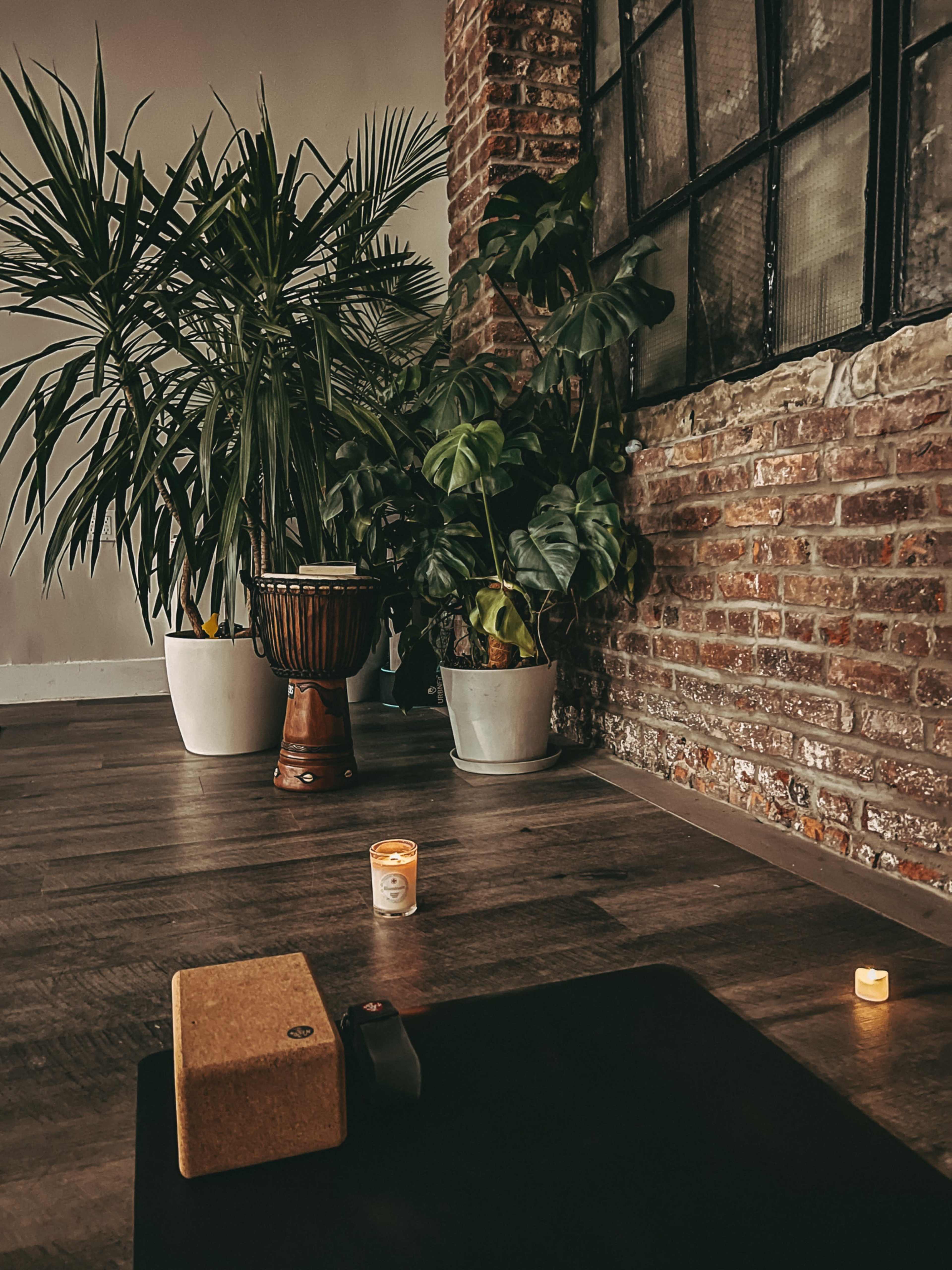 A yoga mat is placed on a wooden floor in front of potted plants and a candle near a large window with a brick wall backdrop.