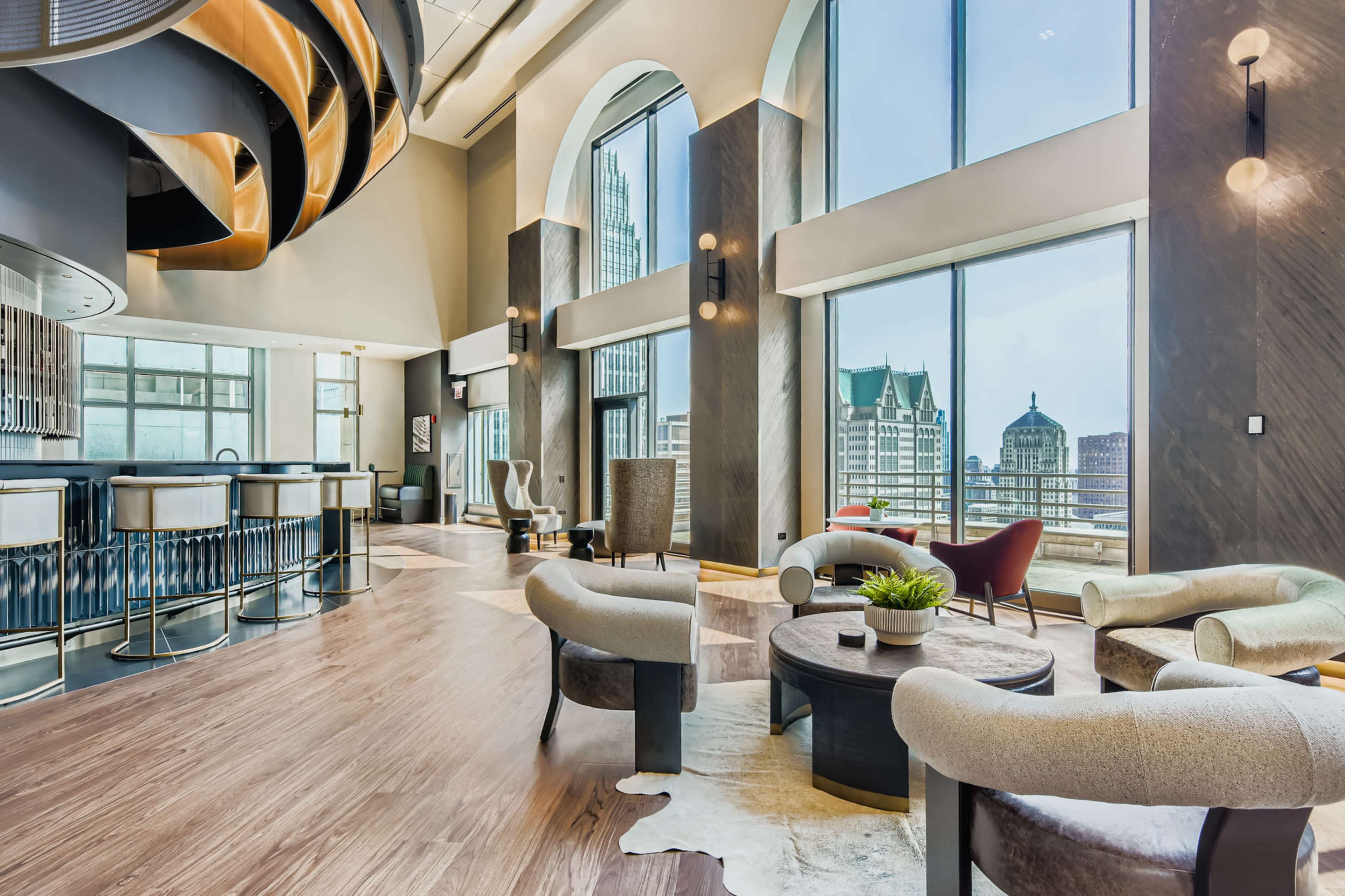 The image shows a modern lounge area with large windows, contemporary furniture, and a stylish bar, overlooking a city skyline.
