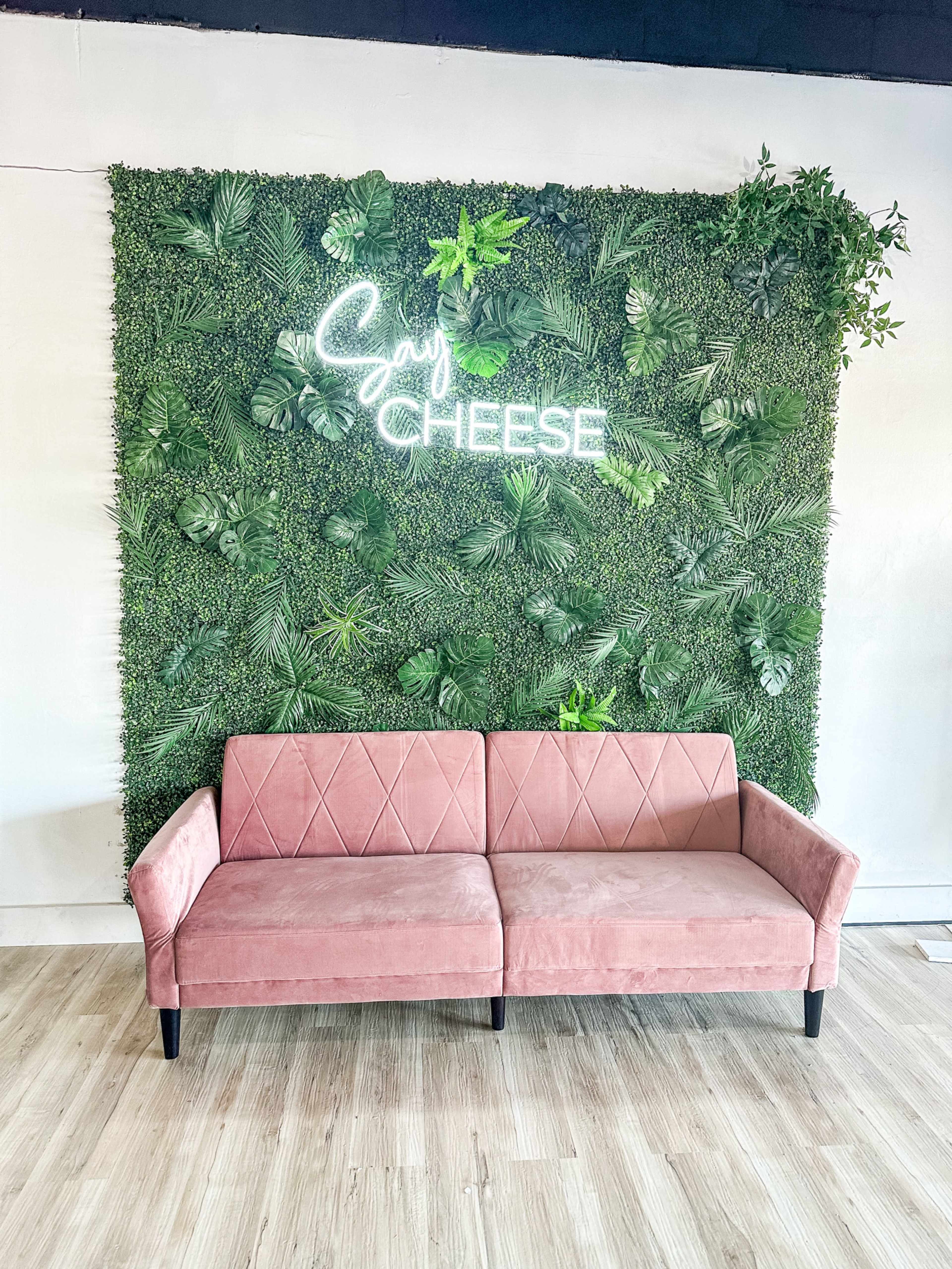 A pink, tufted sofa is placed in front of a green, leafy wall with the words "Say Cheese" illuminated in neon.