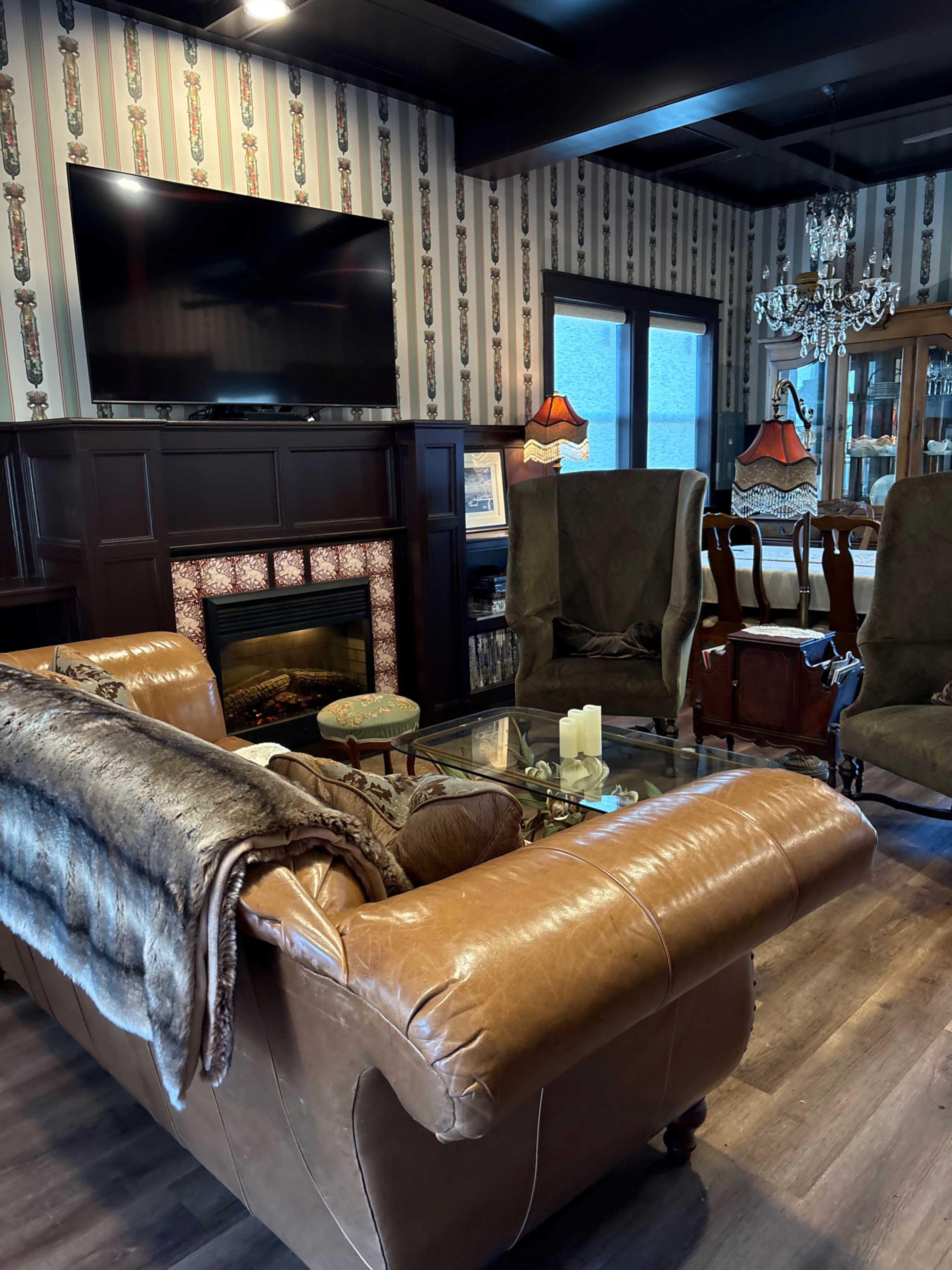 The image shows a cozy living room with a large leather sofa, two green armchairs, a glass coffee table, a fireplace, a mounted television, and a dining area visible in the background.