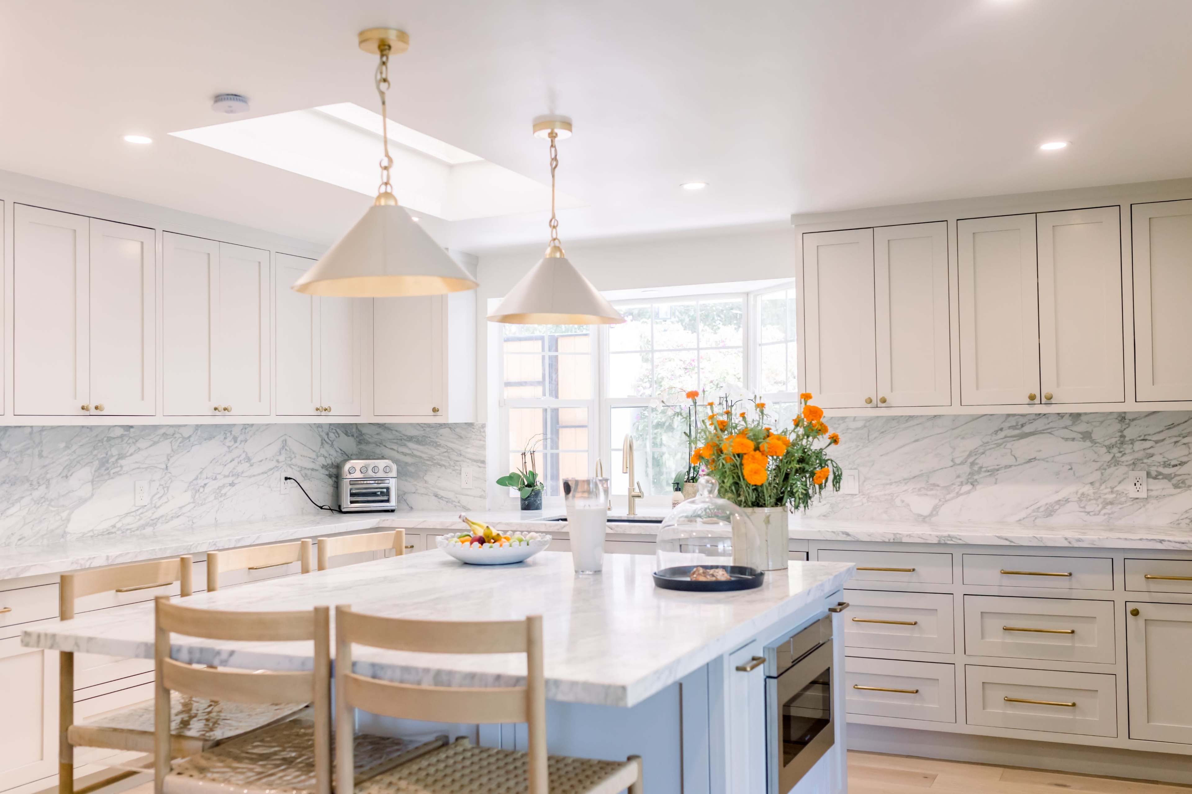 A modern kitchen featuring white cabinets, a marble countertop island, and a large window with a bouquet of orange flowers on the counter.