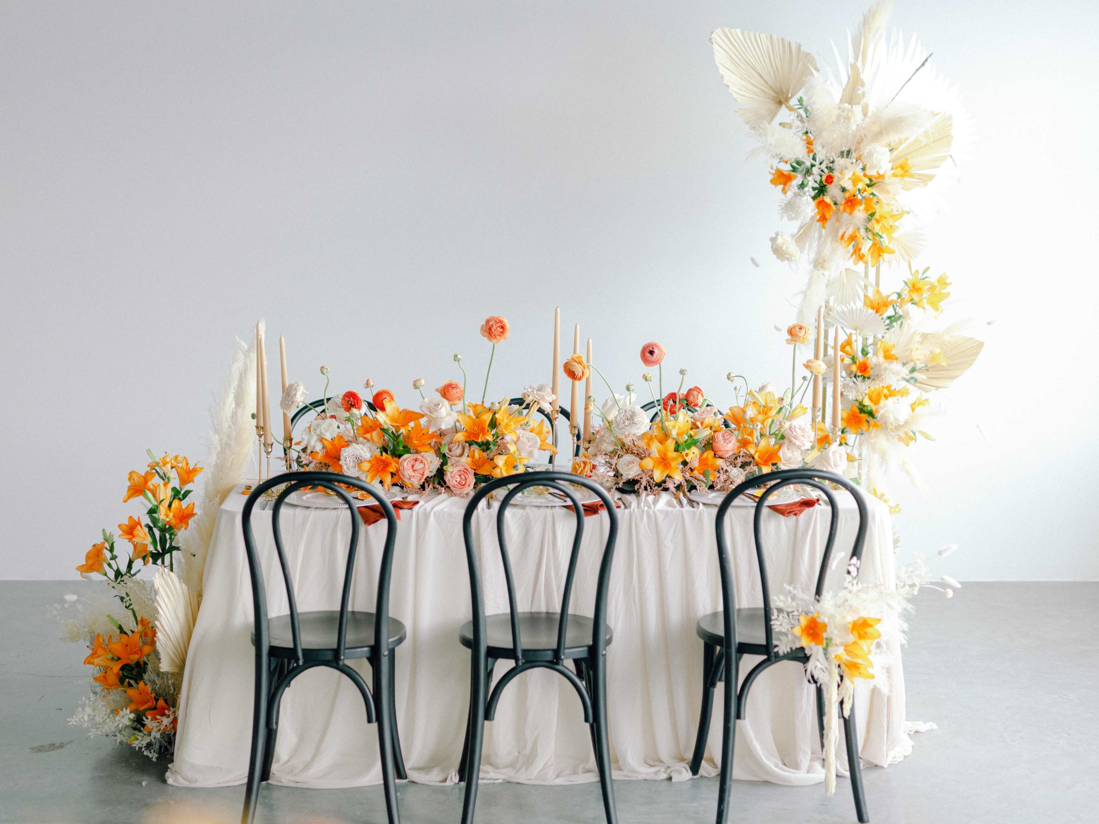 A decorated table with floral arrangements in yellow and orange hues is set with three black chairs facing it.