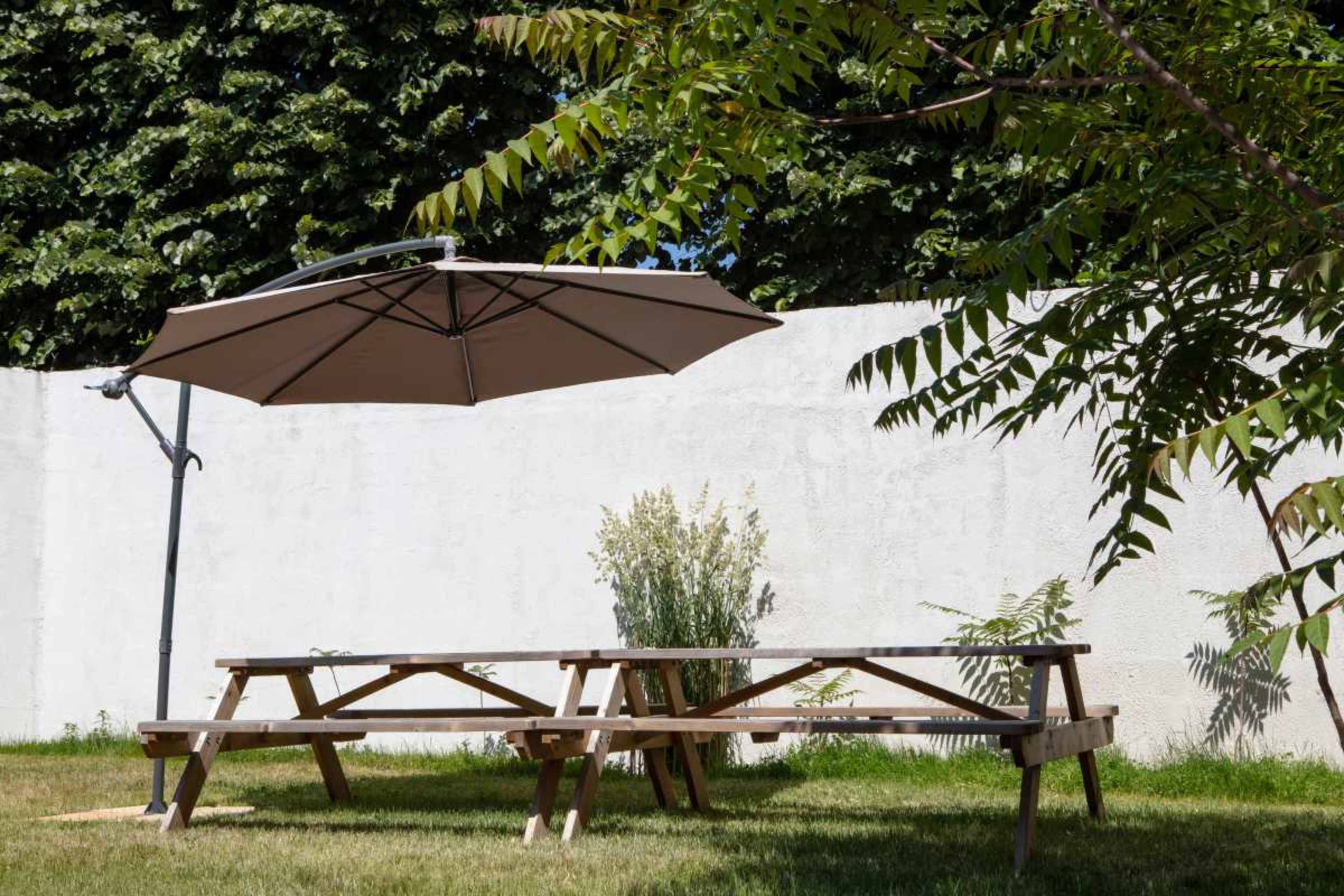 A wooden picnic table with a large umbrella is set on grass beside a white wall and some plants.