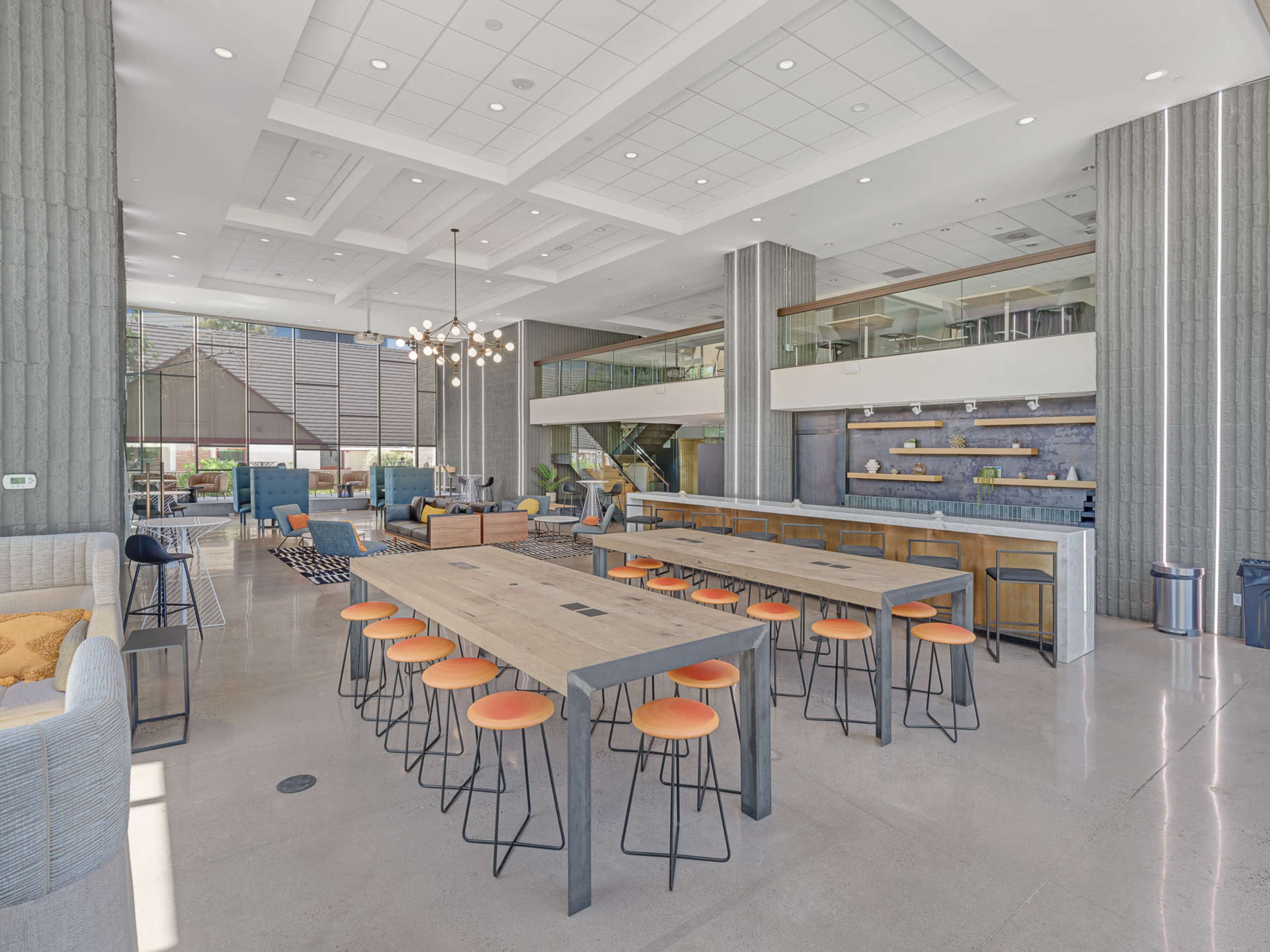 A spacious modern communal area featuring a large dining table with orange stools, a bar area, and various seating arrangements.