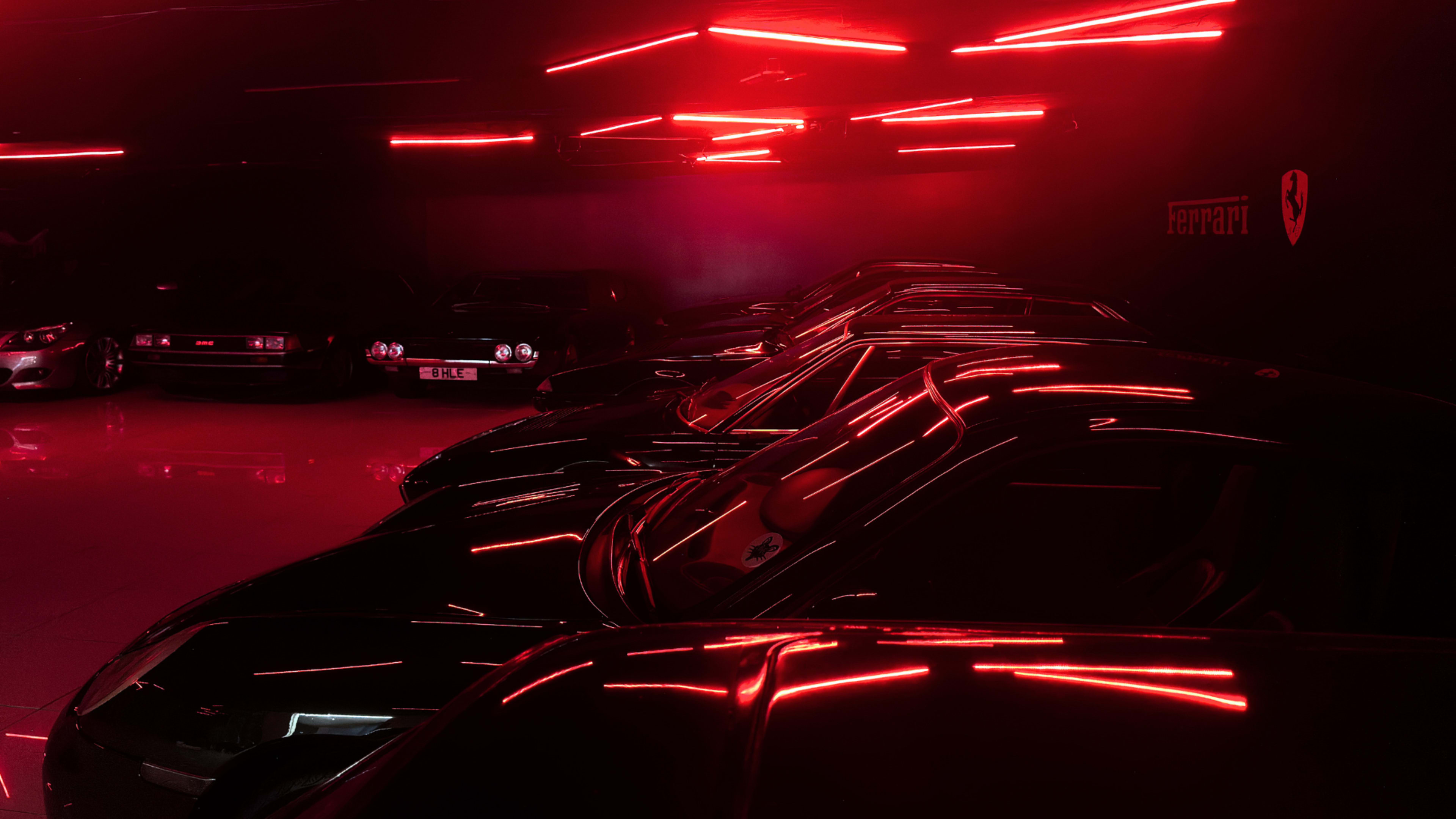 A row of black cars is illuminated by red neon lights in a dark garage.