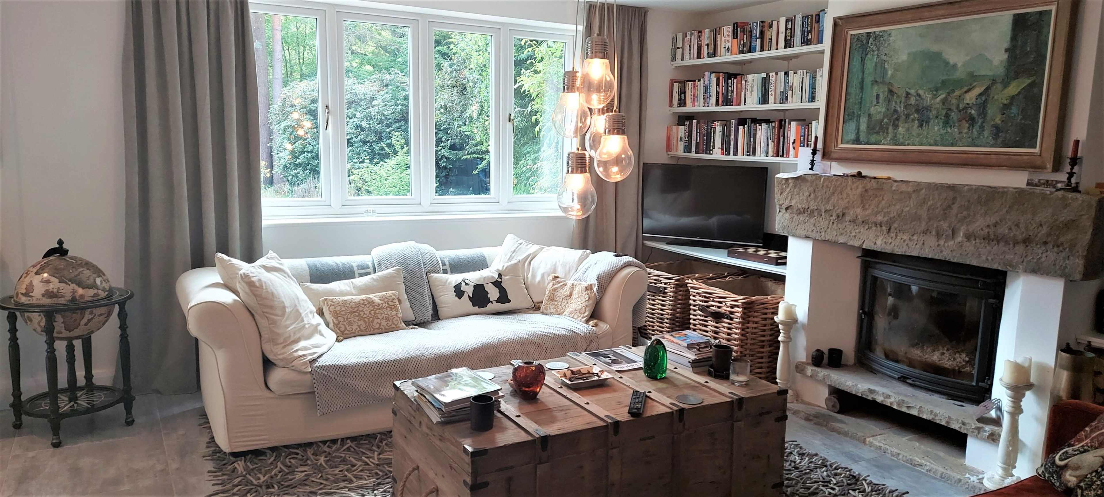 A cozy living room features a white sofa, a wooden coffee table, a stone fireplace, and large windows overlooking greenery.