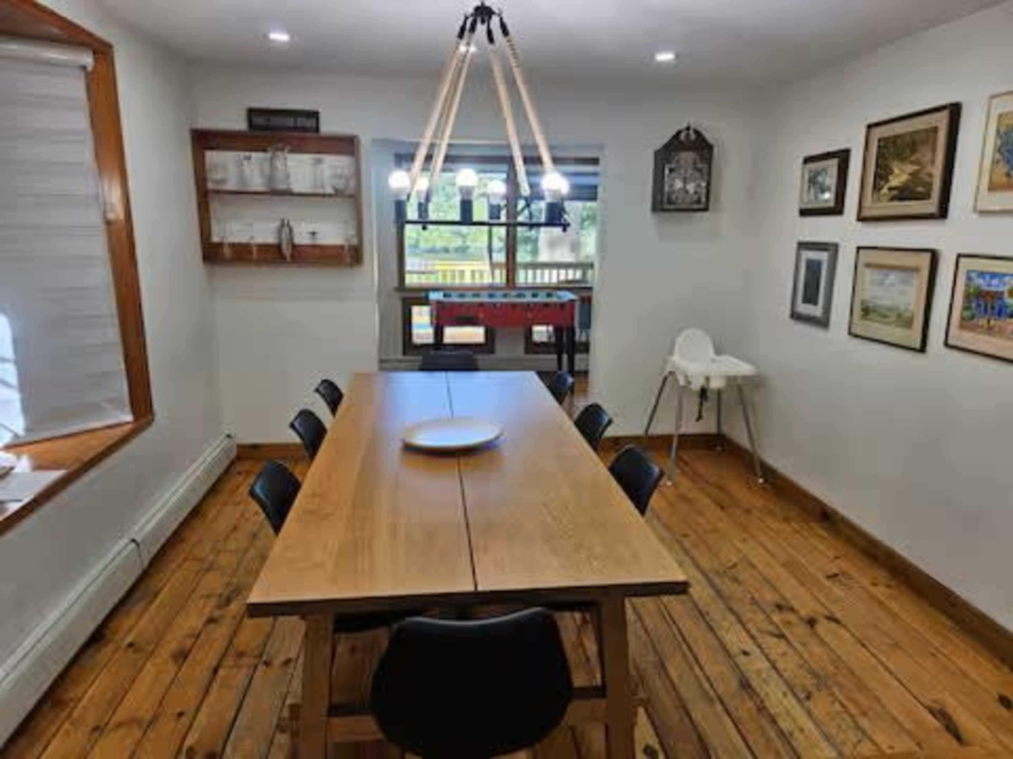 A long wooden dining table with black chairs is placed in a well-lit room featuring a window, a shelf, and several framed pictures on the walls.
