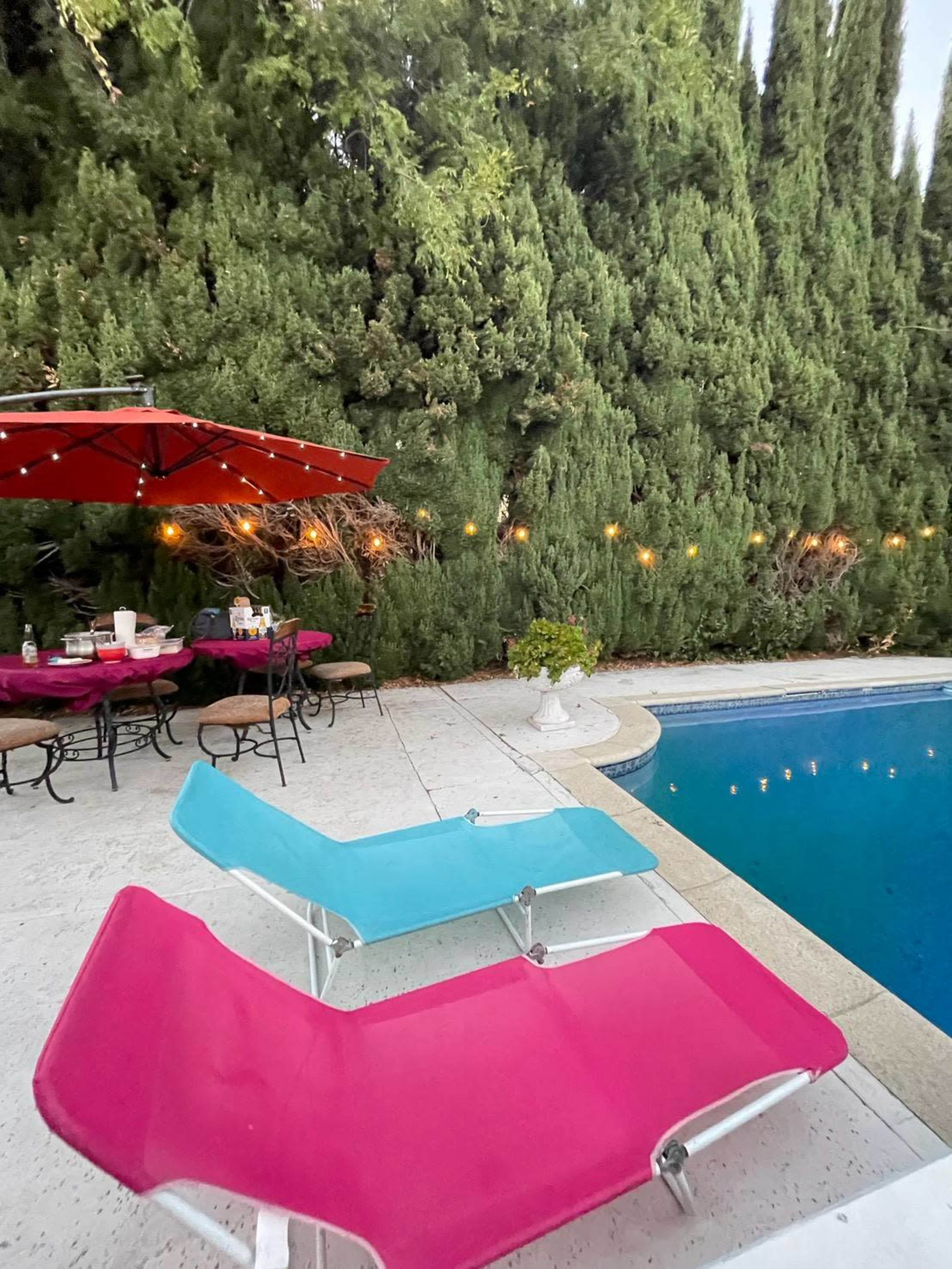 A pink and blue lounge chair are positioned beside a swimming pool, with a table set under an umbrella and greenery lining the background.