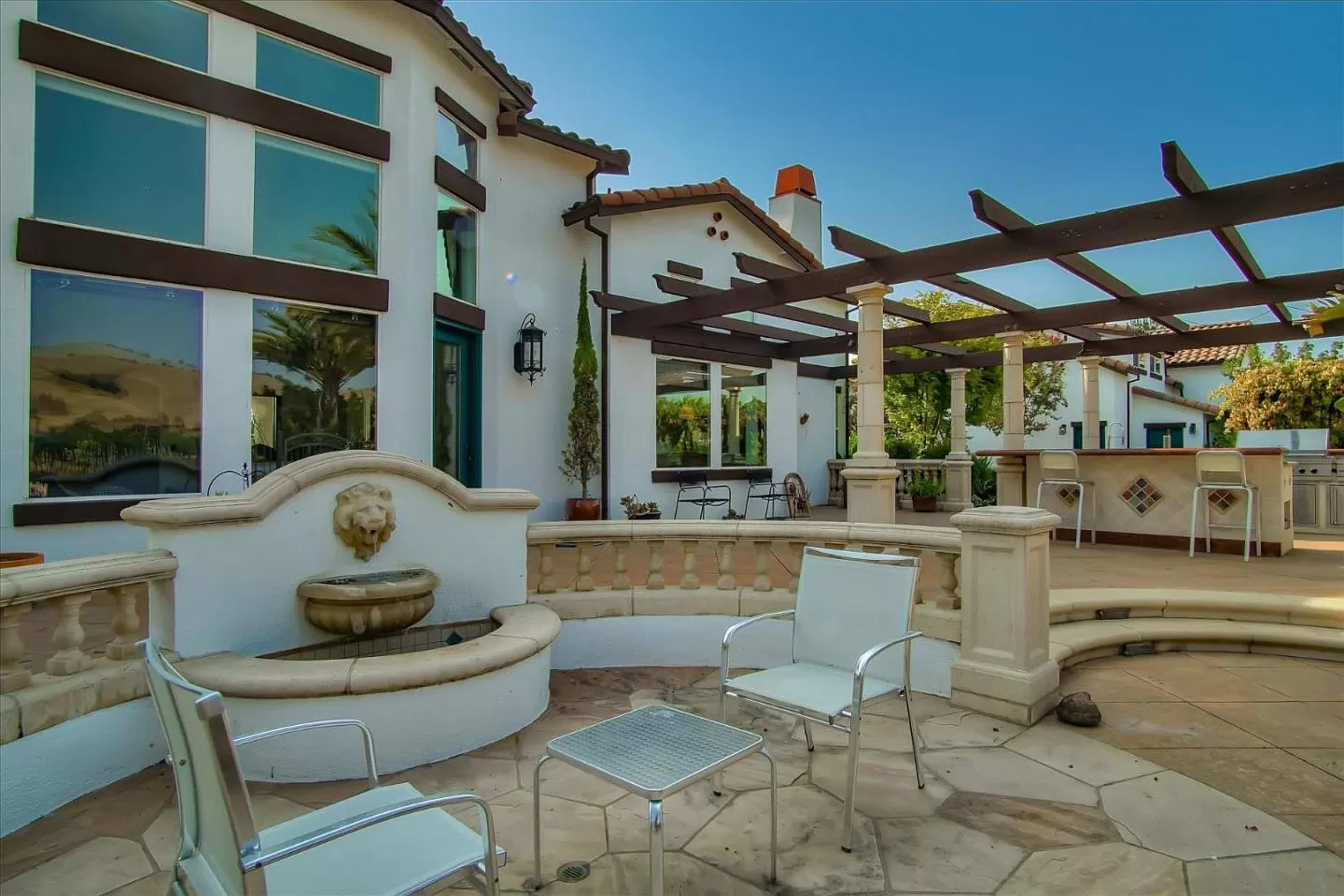 A landscaped outdoor patio area with a fountain, seating chairs, and a pergola near a residential building.