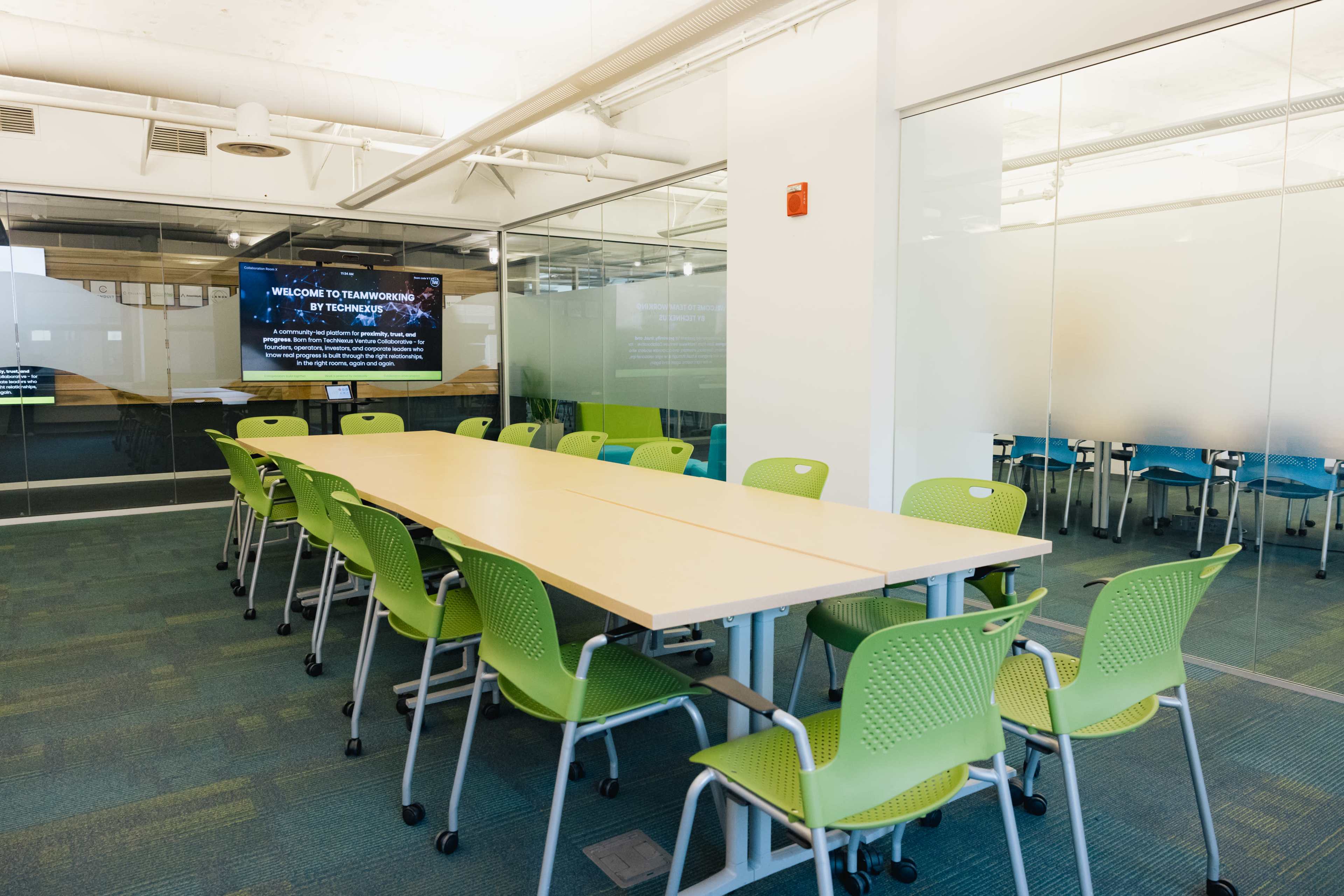 A large, modern conference room features a long wooden table surrounded by green chairs, with large glass walls displaying a digital welcome message.