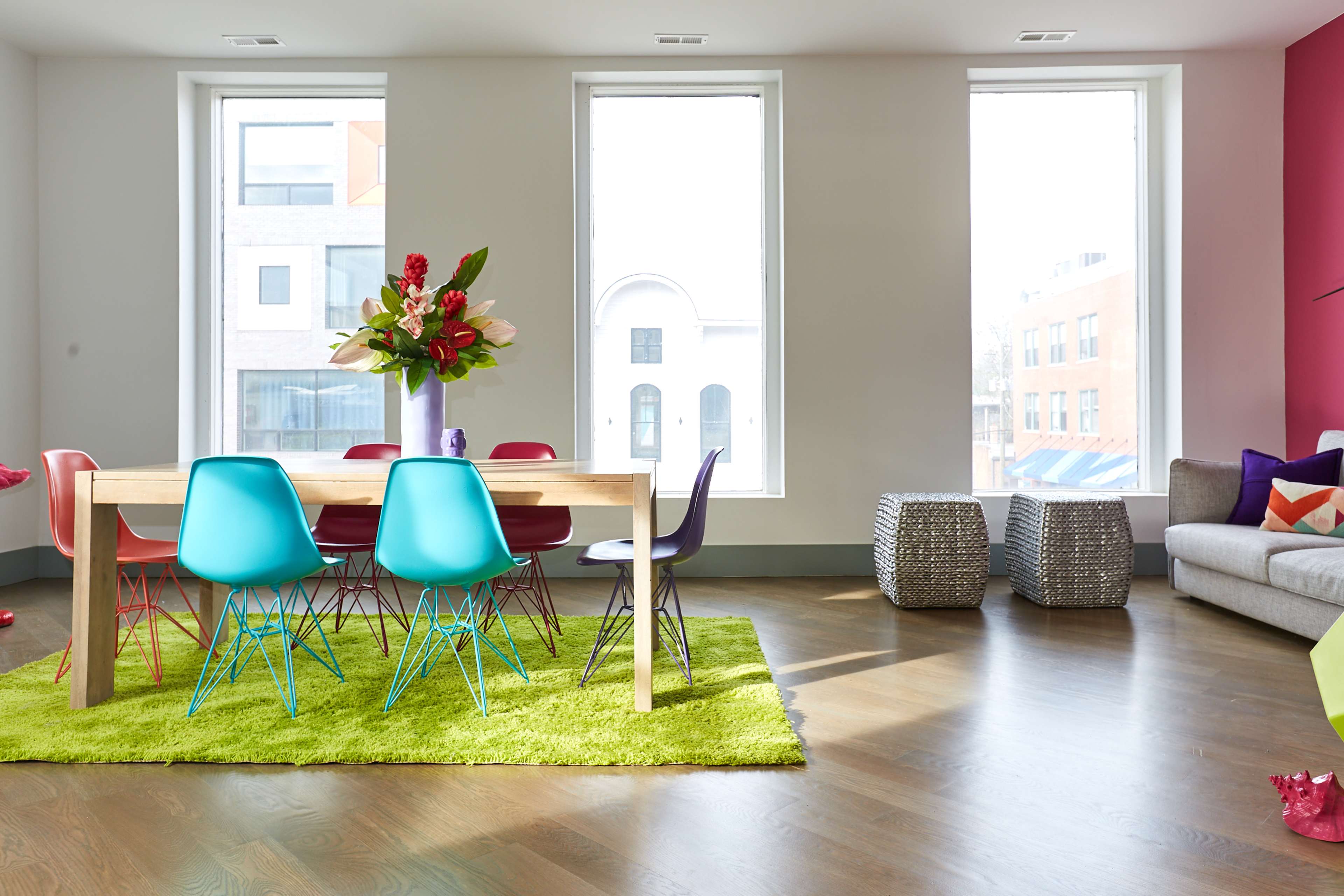 A modern dining area features a wooden table surrounded by colorful chairs on a green rug, with large windows allowing natural light to fill the space.