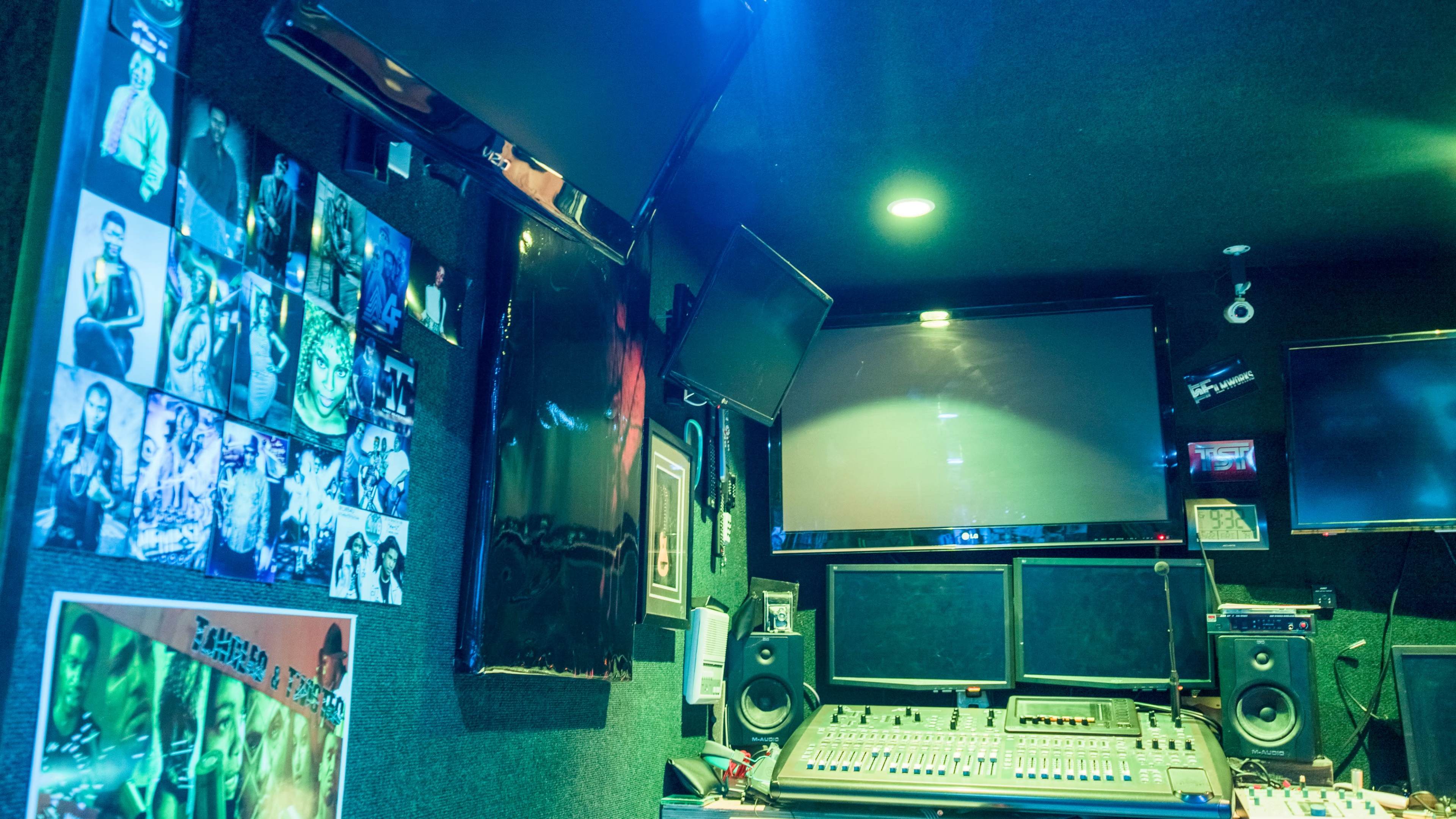 A recording studio with multiple screens, a sound mixing console, and framed images on the walls.
