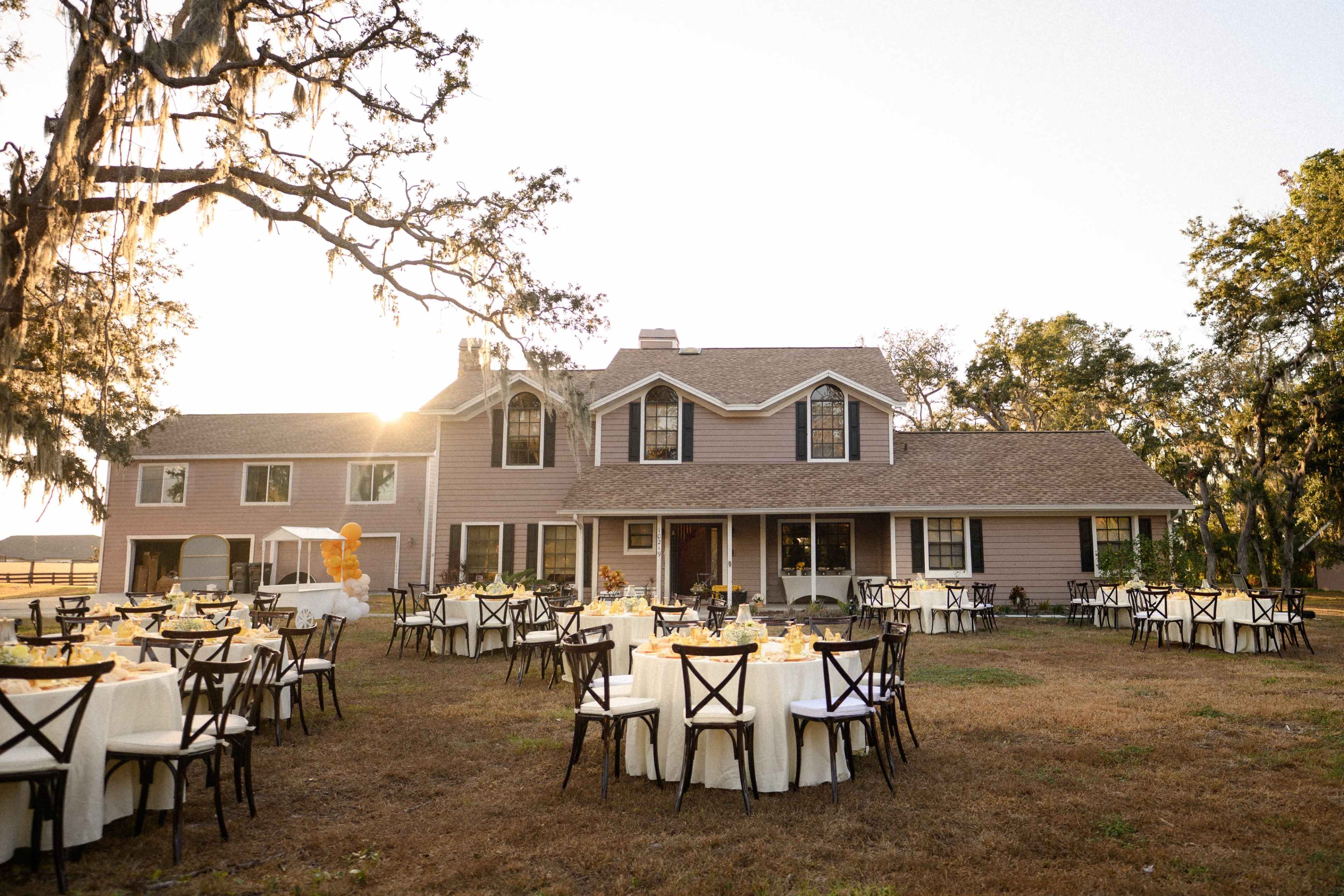 A large house with a sloped roof is surrounded by tables set for an outdoor event in a grassy area.