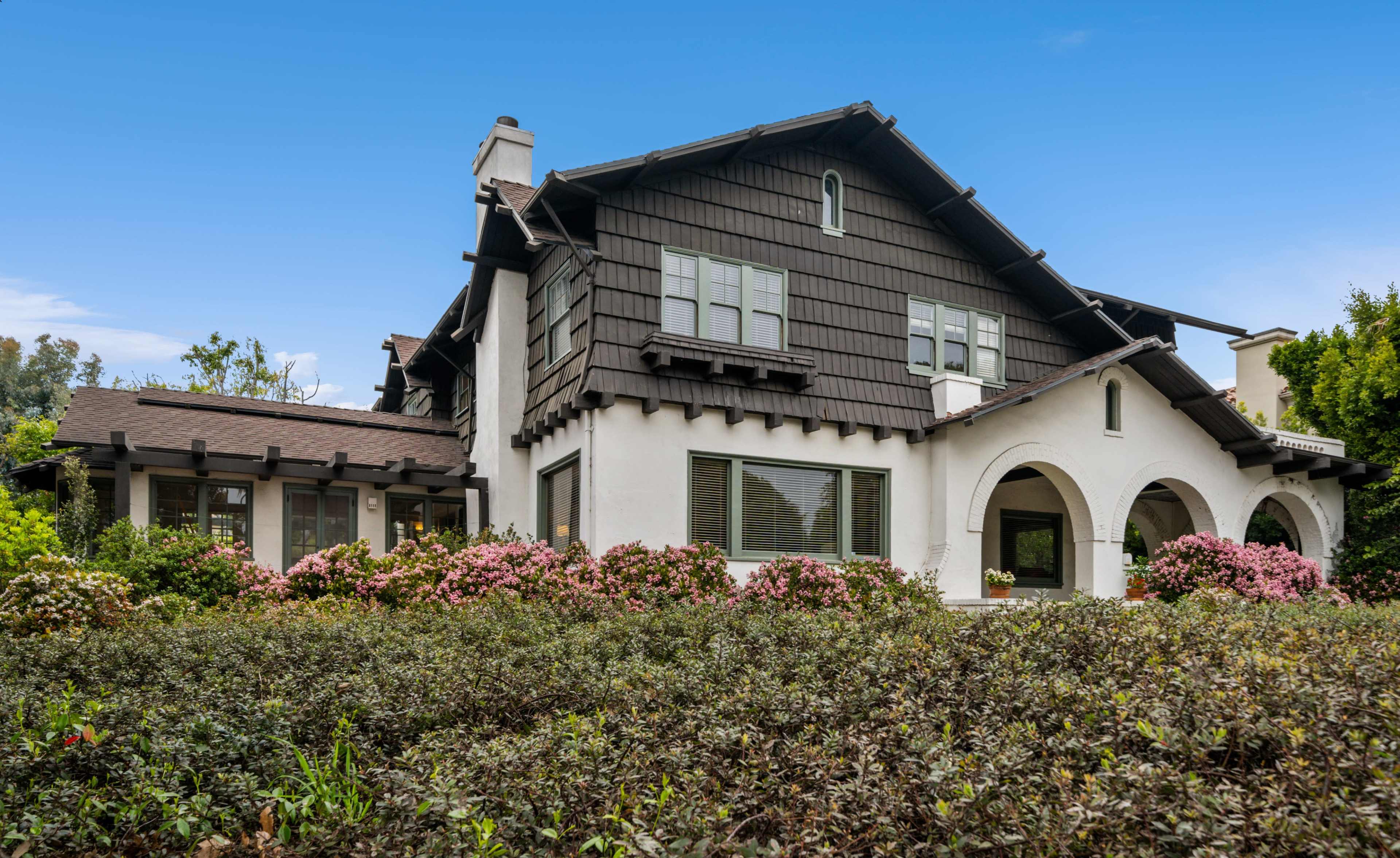 A two-story house with a dark brown shingle exterior and a white stucco lower facade is surrounded by flowering shrubs and greenery.