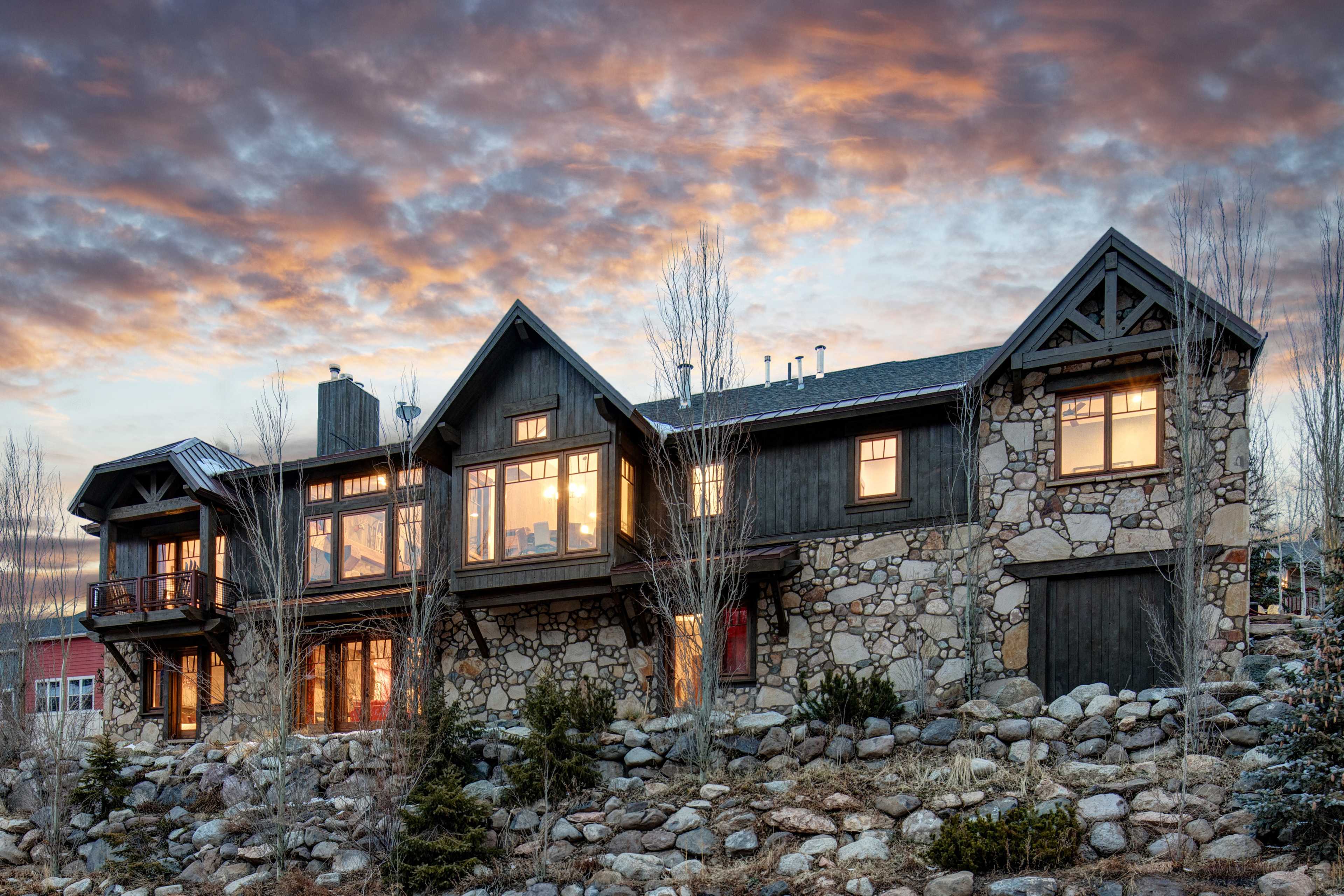 A large stone and wooden house with numerous windows sits on a rocky hillside under a dramatic sunset sky.