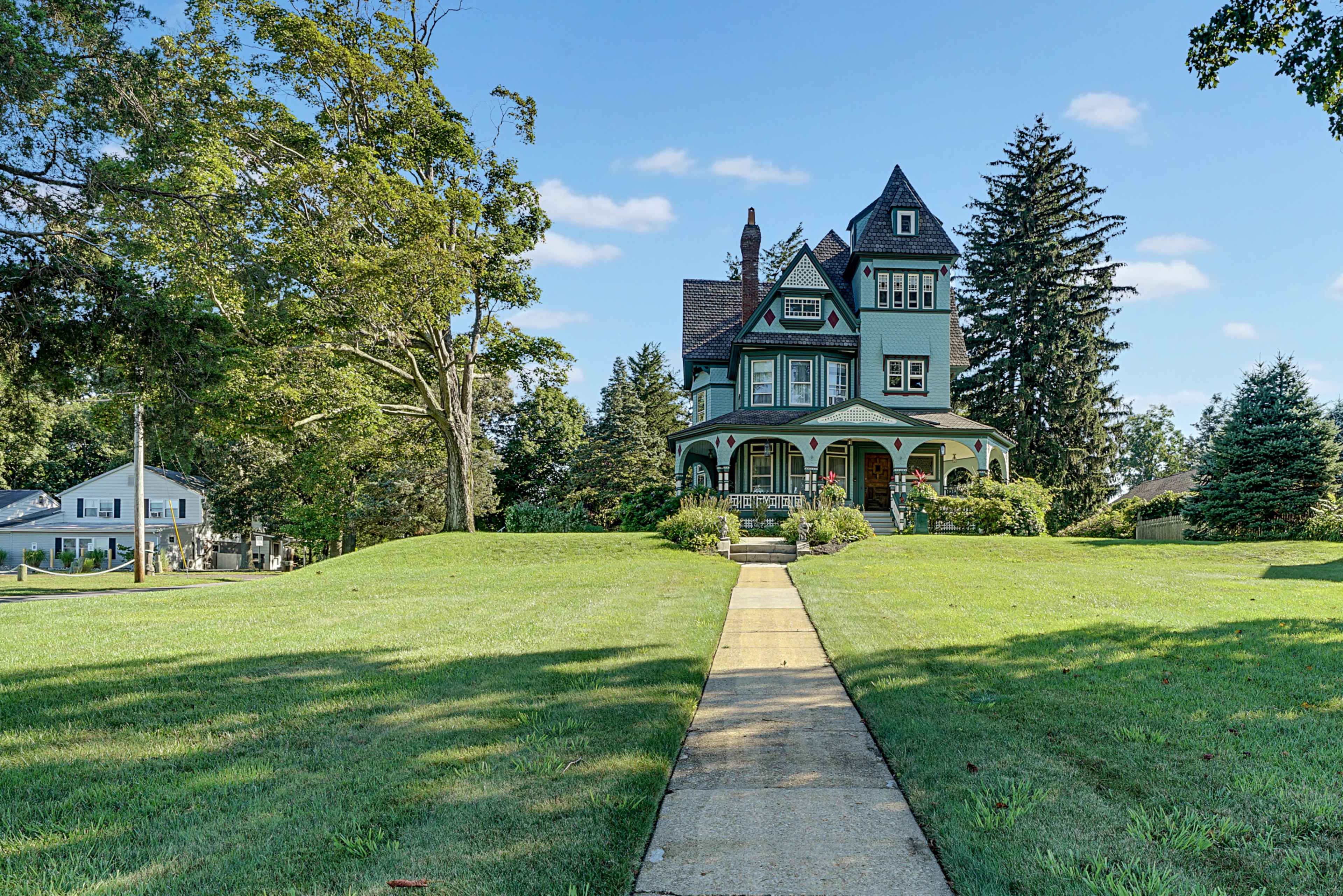 A large, green Victorian-style house stands at the end of a concrete path, surrounded by trees and a manicured lawn.