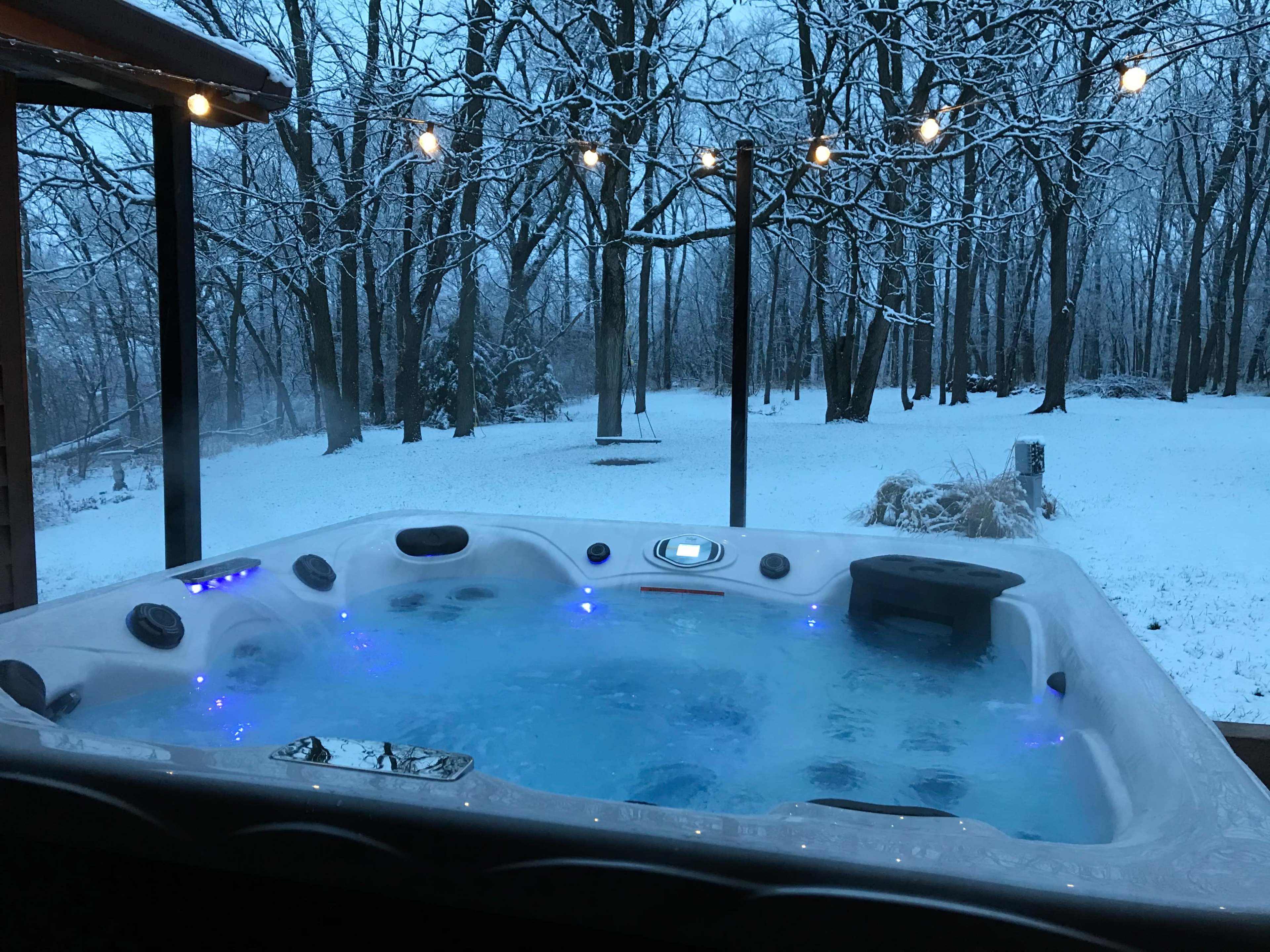 A hot tub filled with water and blue lights is positioned on a deck, overlooking a snowy landscape with trees in the background.