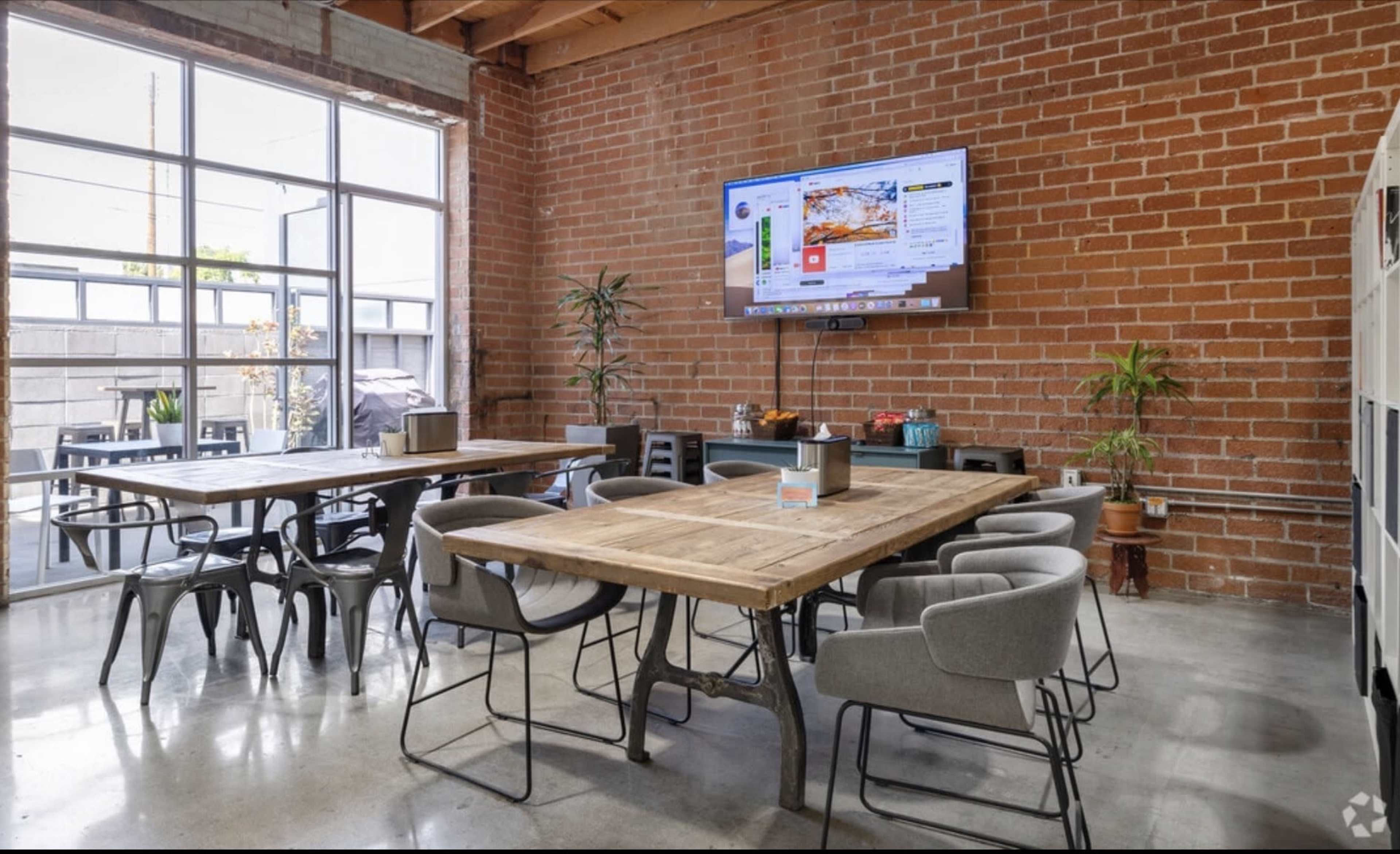 The image shows a spacious, modern meeting room with brick walls, wooden tables, and a large wall-mounted television.