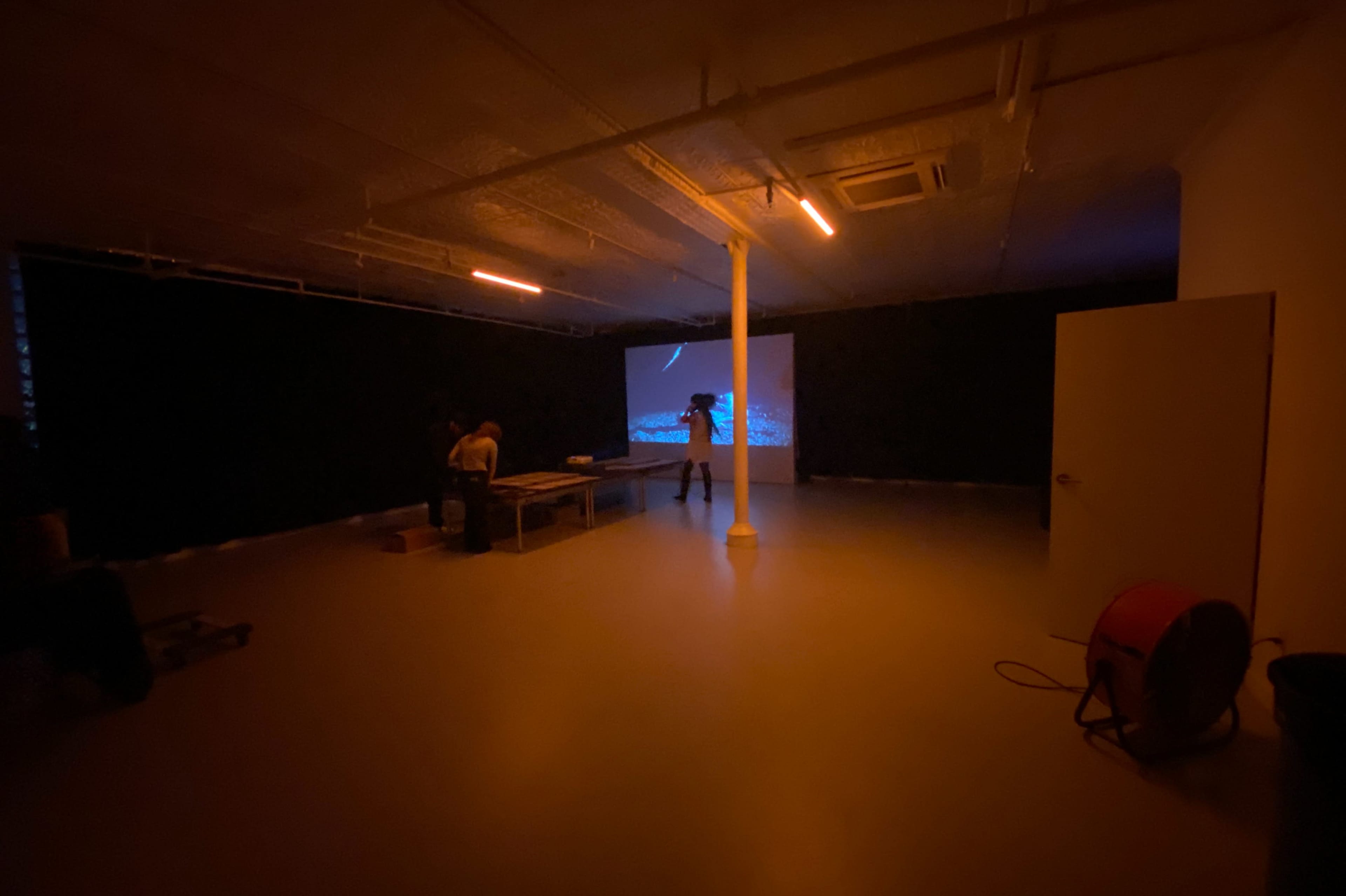A dimly lit room with two people working at a table while a projection illuminates a wall.