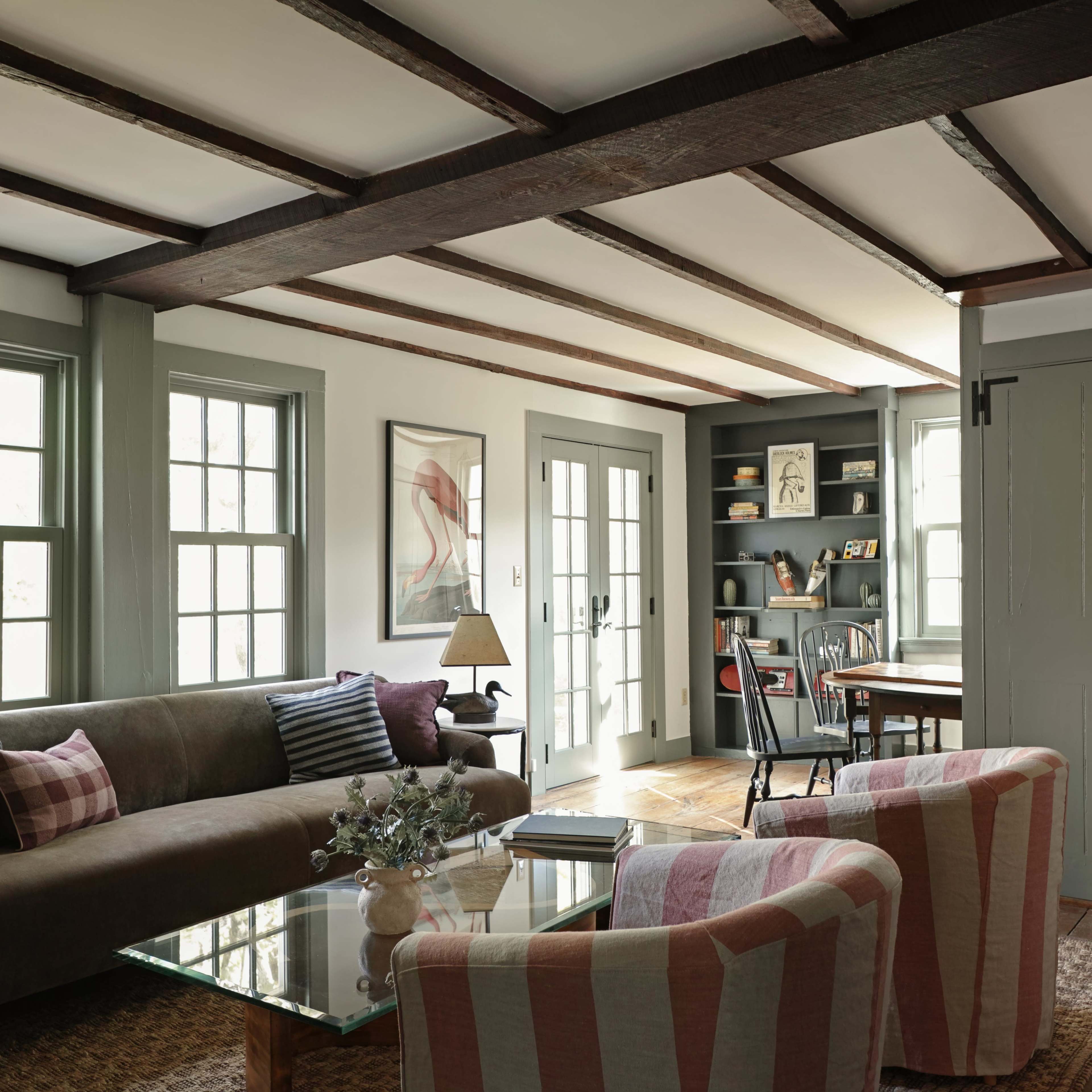 The image shows a cozy living room featuring a brown couch, two striped armchairs, a glass coffee table, and a desk in a corner with shelves filled with books.