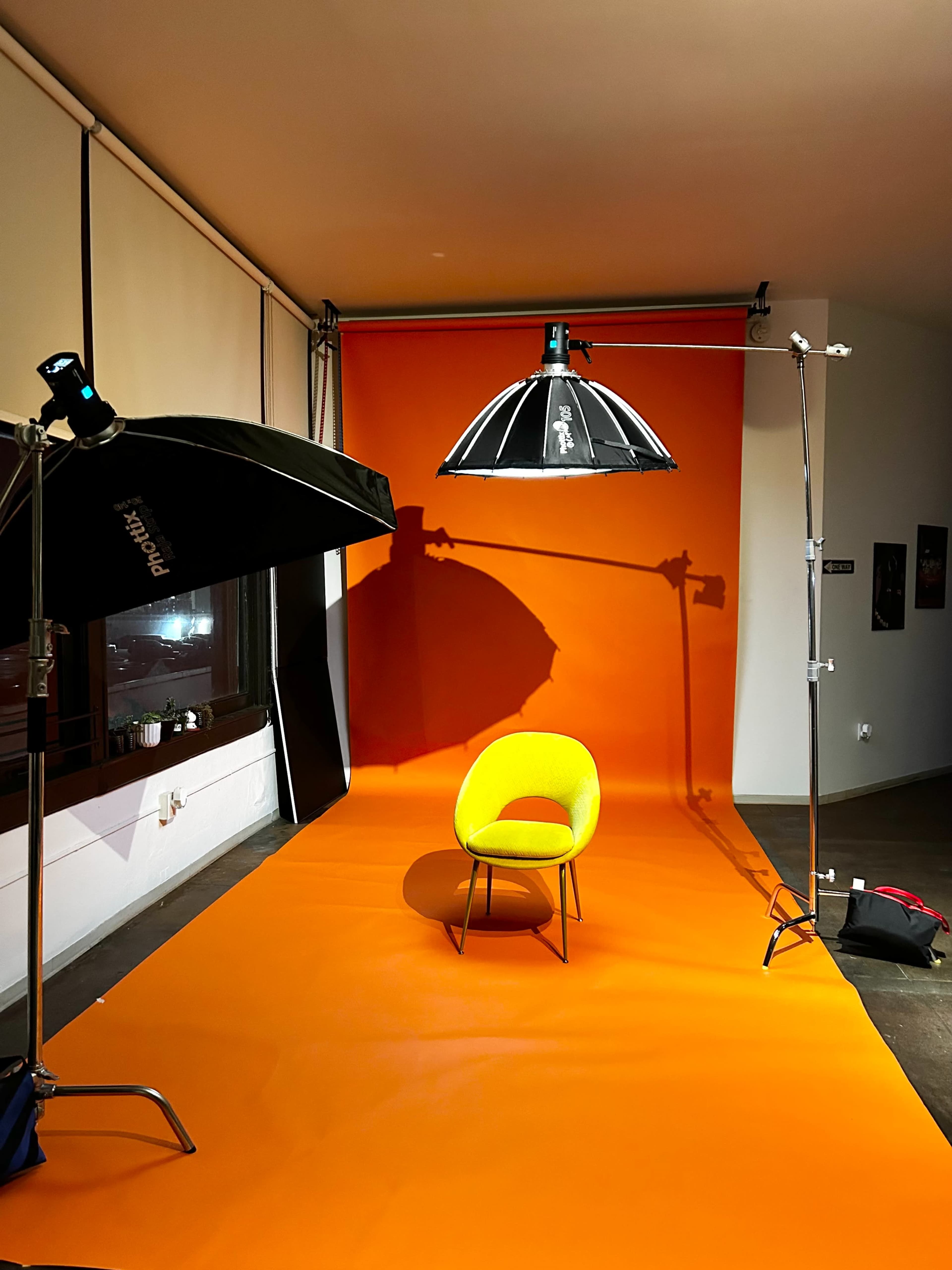 The image shows a photography studio with an orange backdrop, a chair in the center, and two lighting setups.