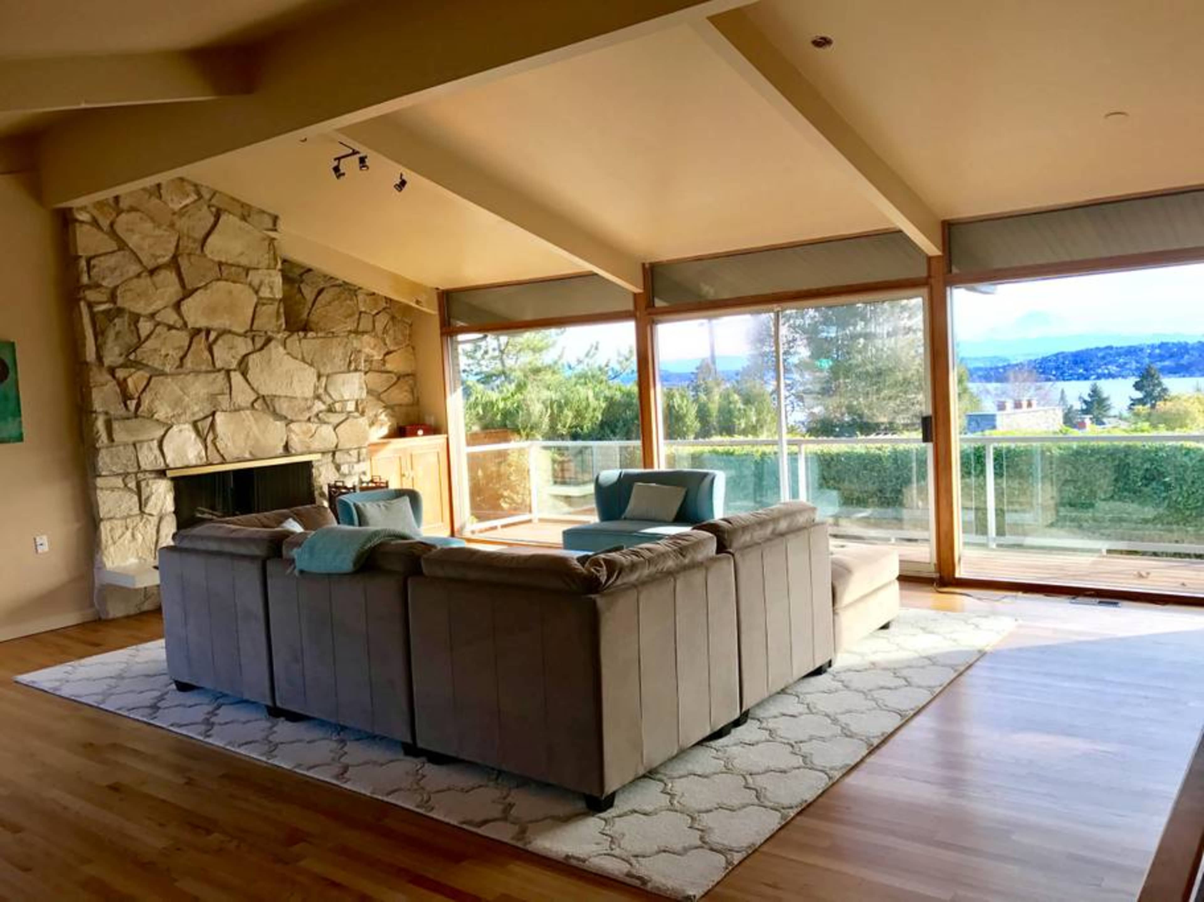 A spacious living room features a stone fireplace, large windows with views of a landscape, and modern furniture arranged around a light-colored area rug.