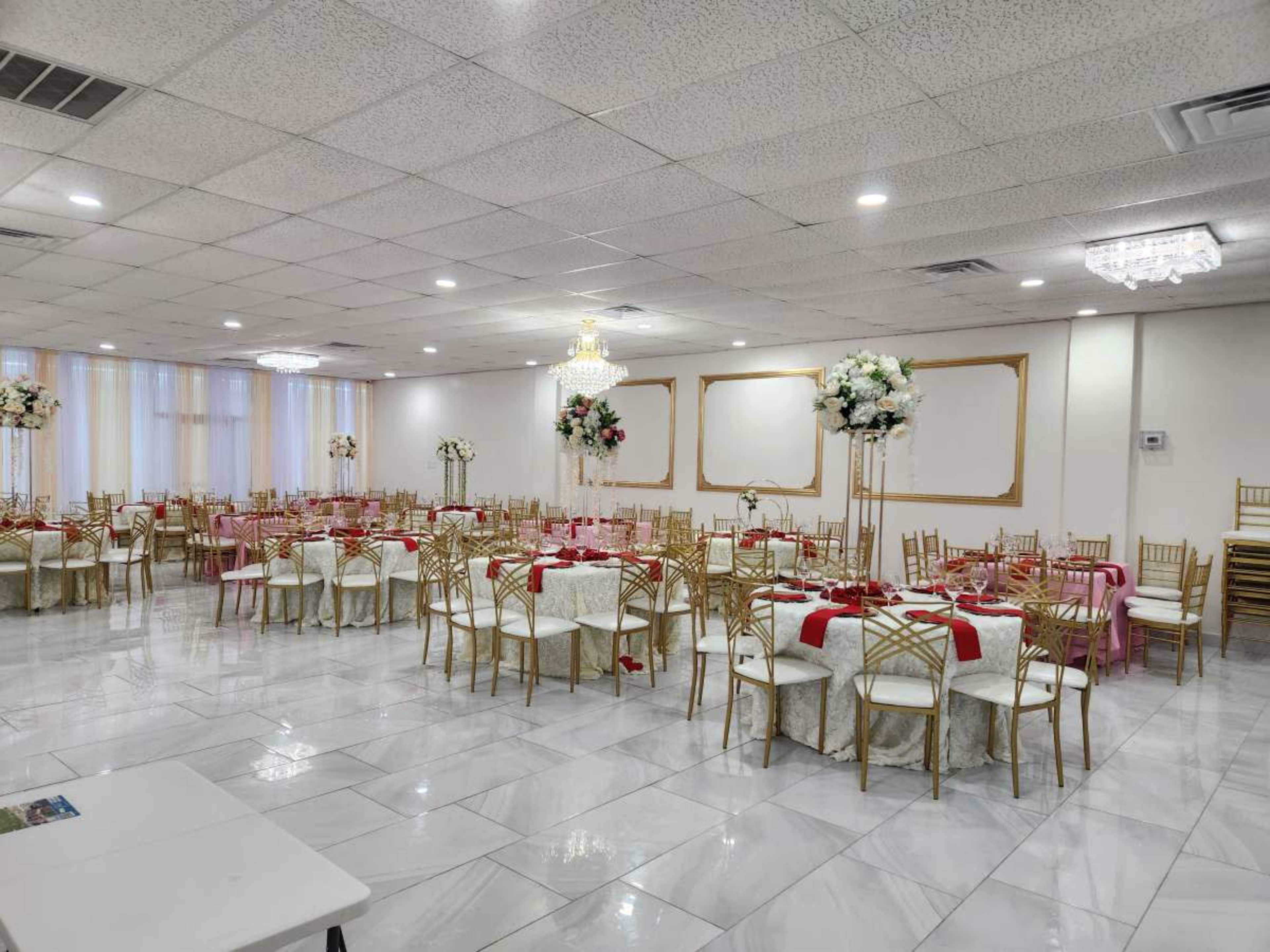 A banquet hall is set up with round tables covered in white tablecloths and gold-framed chairs, decorated with red accents and floral arrangements.