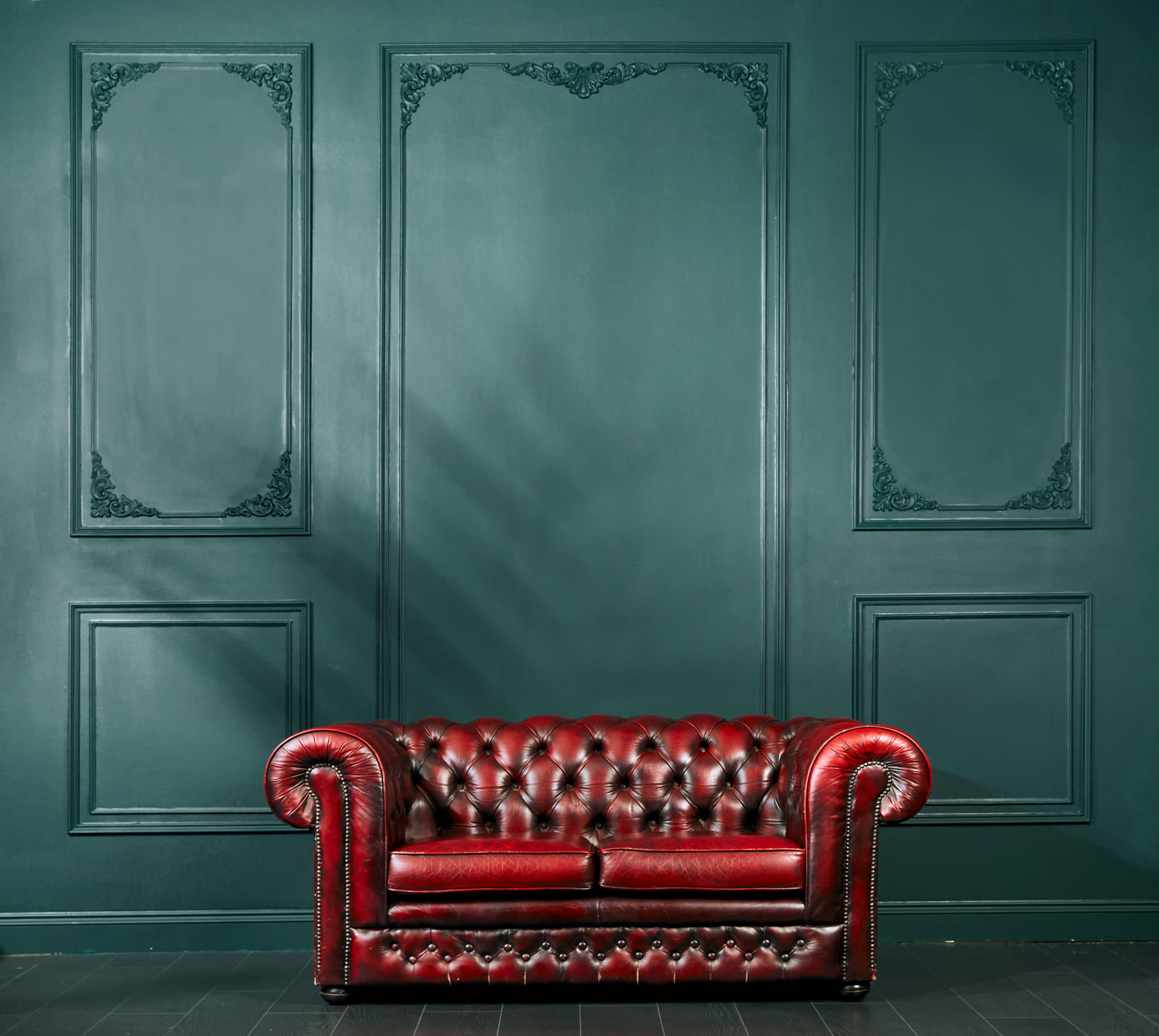A red leather Chesterfield sofa is positioned against a dark green wall with decorative paneling.