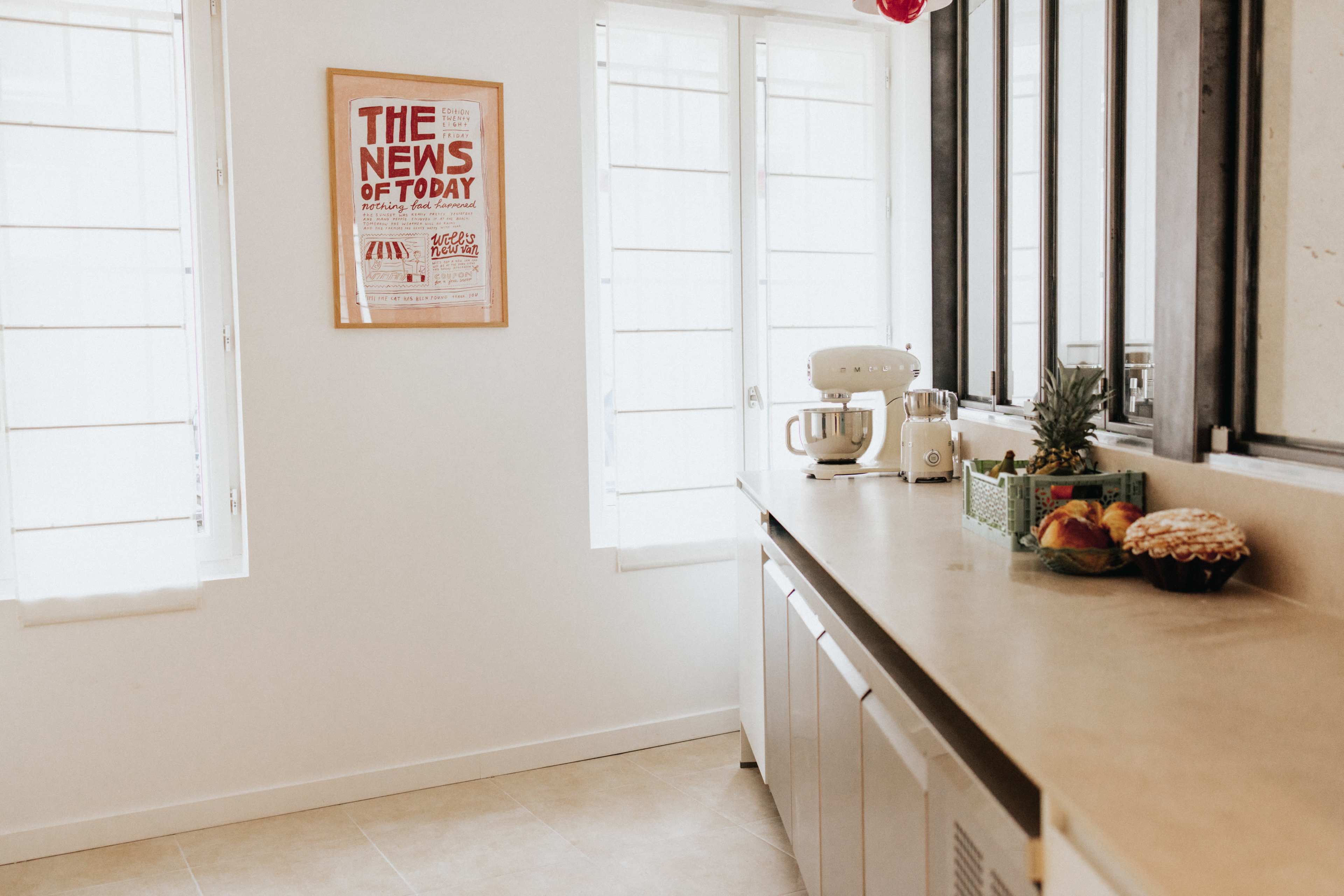 A modern kitchen features a countertop with a stand mixer, a basket of fruits, and a decorative poster on the wall, with sunlight streaming through the windows.