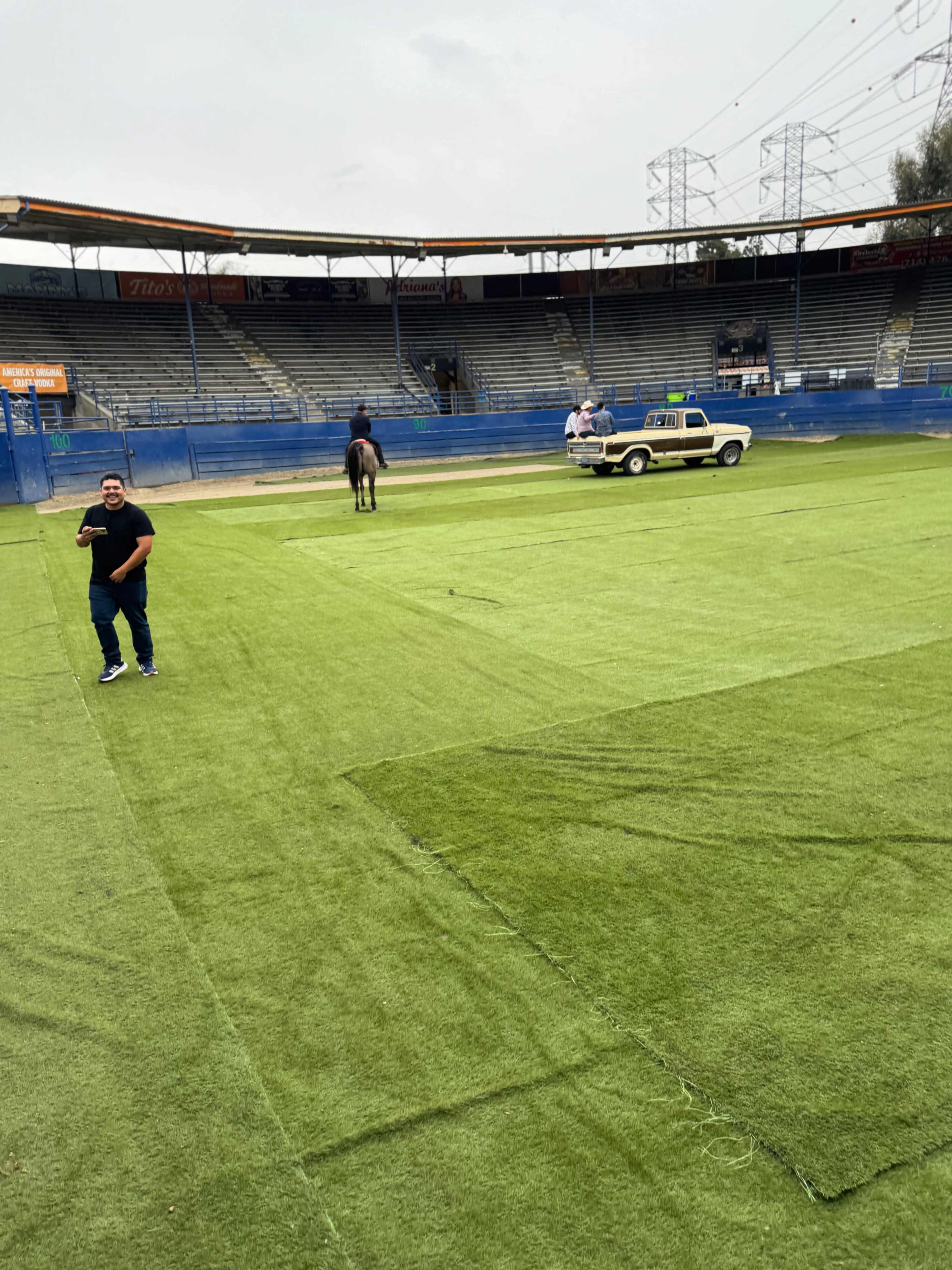 A group of people is installing artificial turf on a sports field, with a truck parked nearby and empty bleachers in the background.