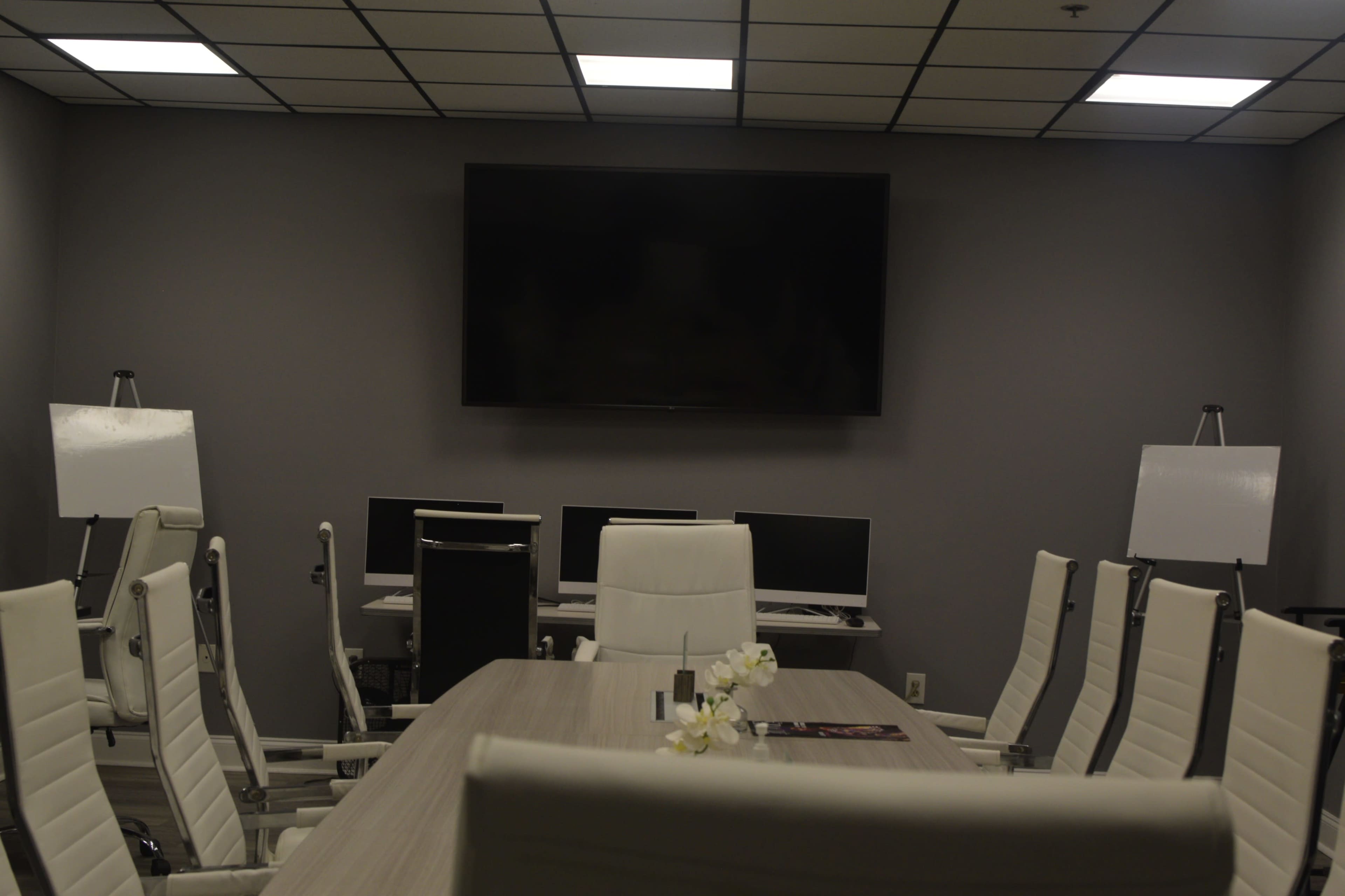 A modern conference room features a long table surrounded by white chairs, a large wall-mounted screen, and several computers on a side desk.